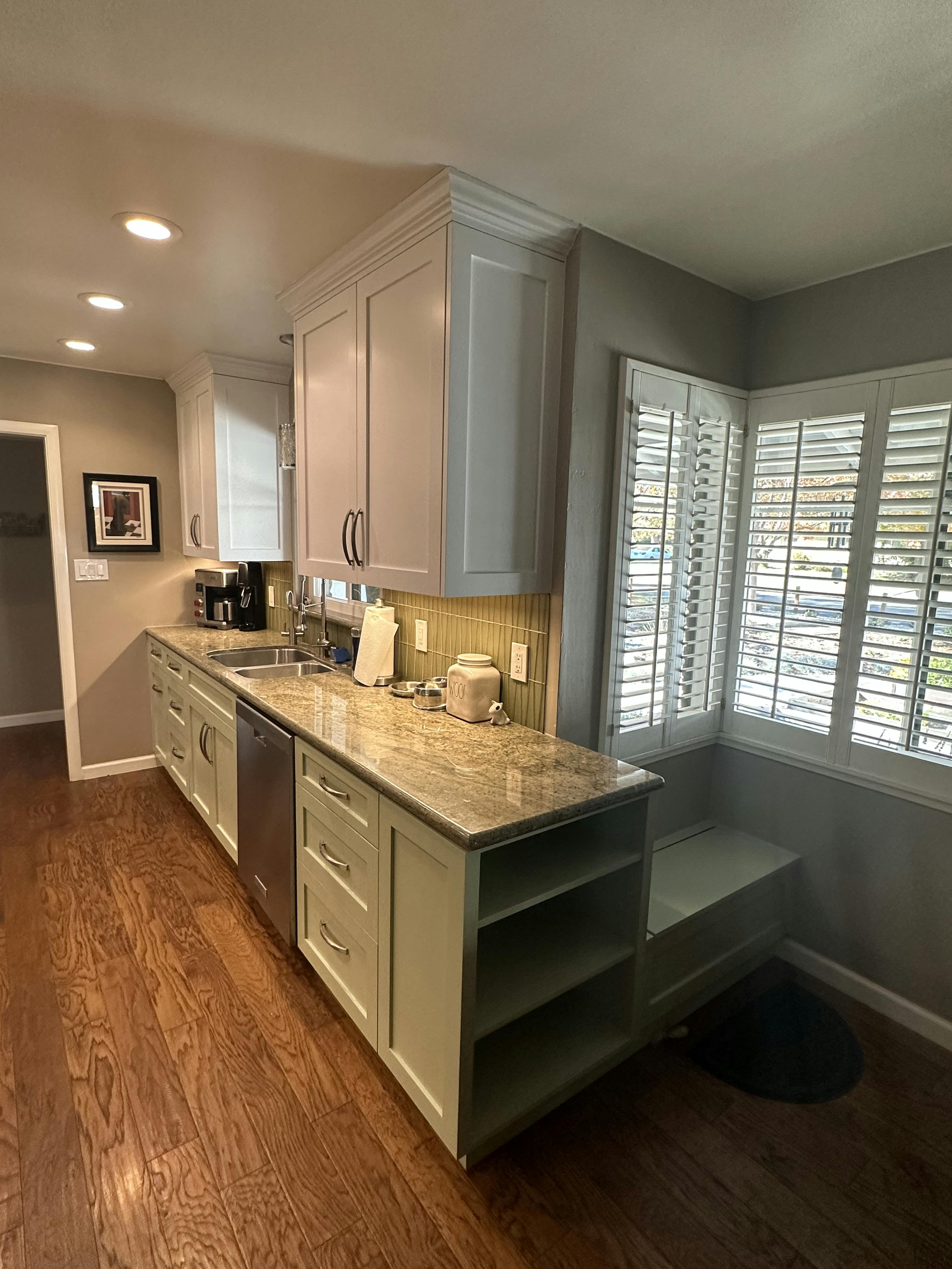 Modern kitchen interior with gray cabinets, beige tile backsplash, and wooden flooring. The space is well-lit with natural light from the windows covered with white shutters.