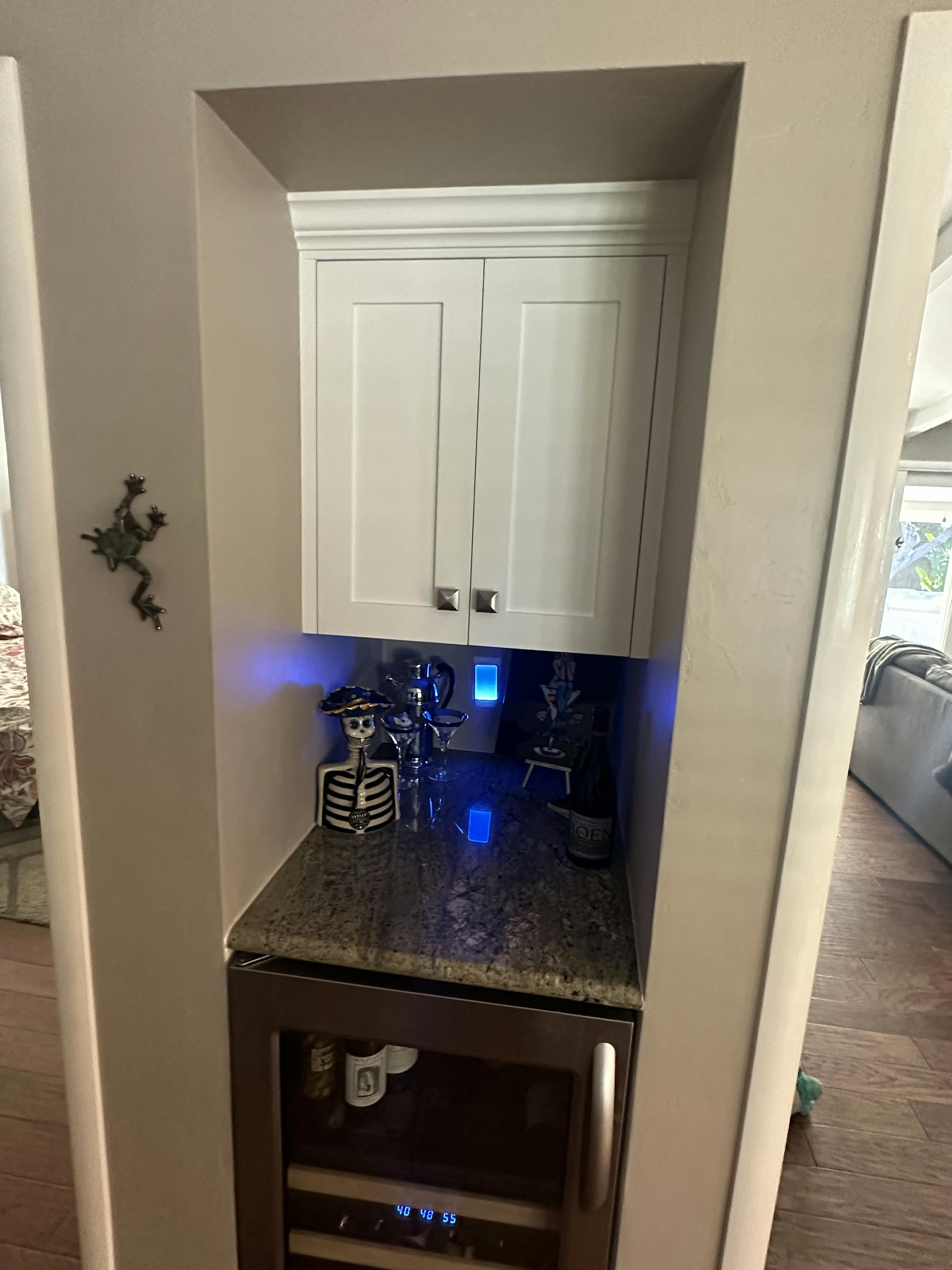 A compact kitchen nook with white cabinets above a granite countertop, a built-in wine cooler below, and decorative items illuminated by blue accent lighting.