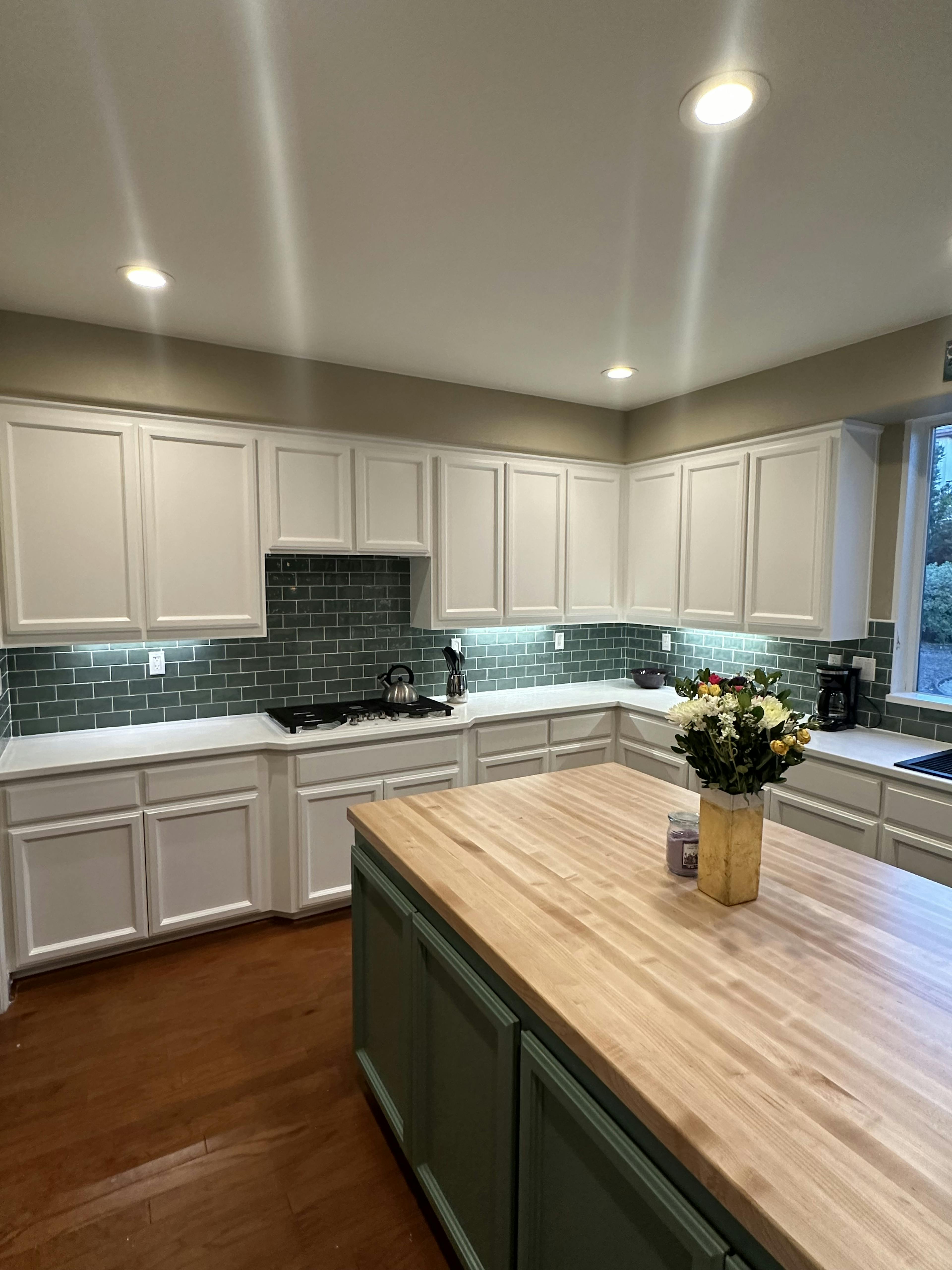 Spacious kitchen interior featuring white cabinetry with panel doors, dark green subway tile backsplash, and wooden flooring. A large wooden island countertop is at the forefront, adorned with a vase of fresh flowers and a candle, under bright ceiling recessed lighting.