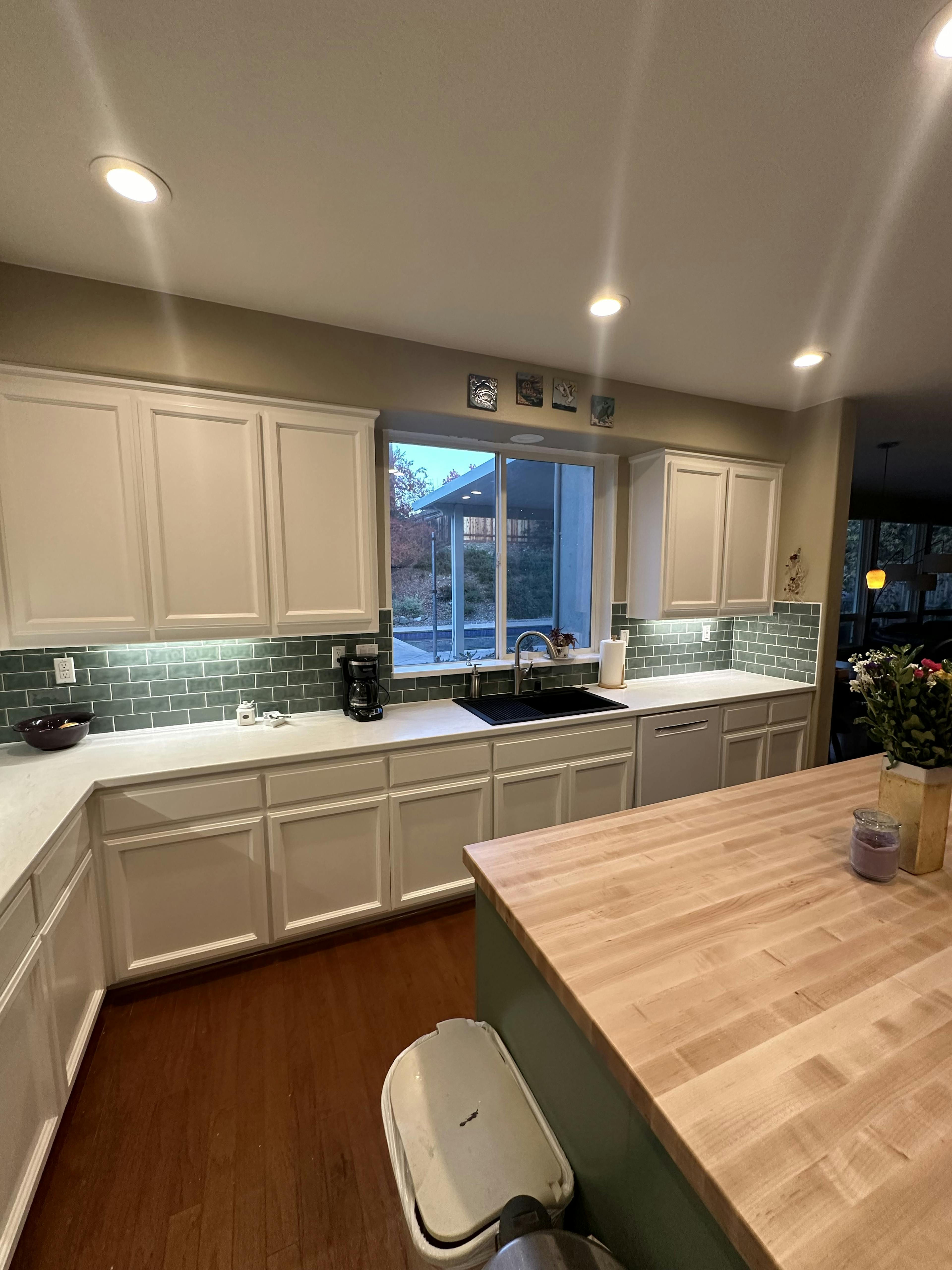 Contemporary kitchen with white cabinetry, dark green subway tile backsplash, and a window overlooking the outside. The wooden floor complements the large wooden island countertop, and the room is illuminated by recessed ceiling lights.