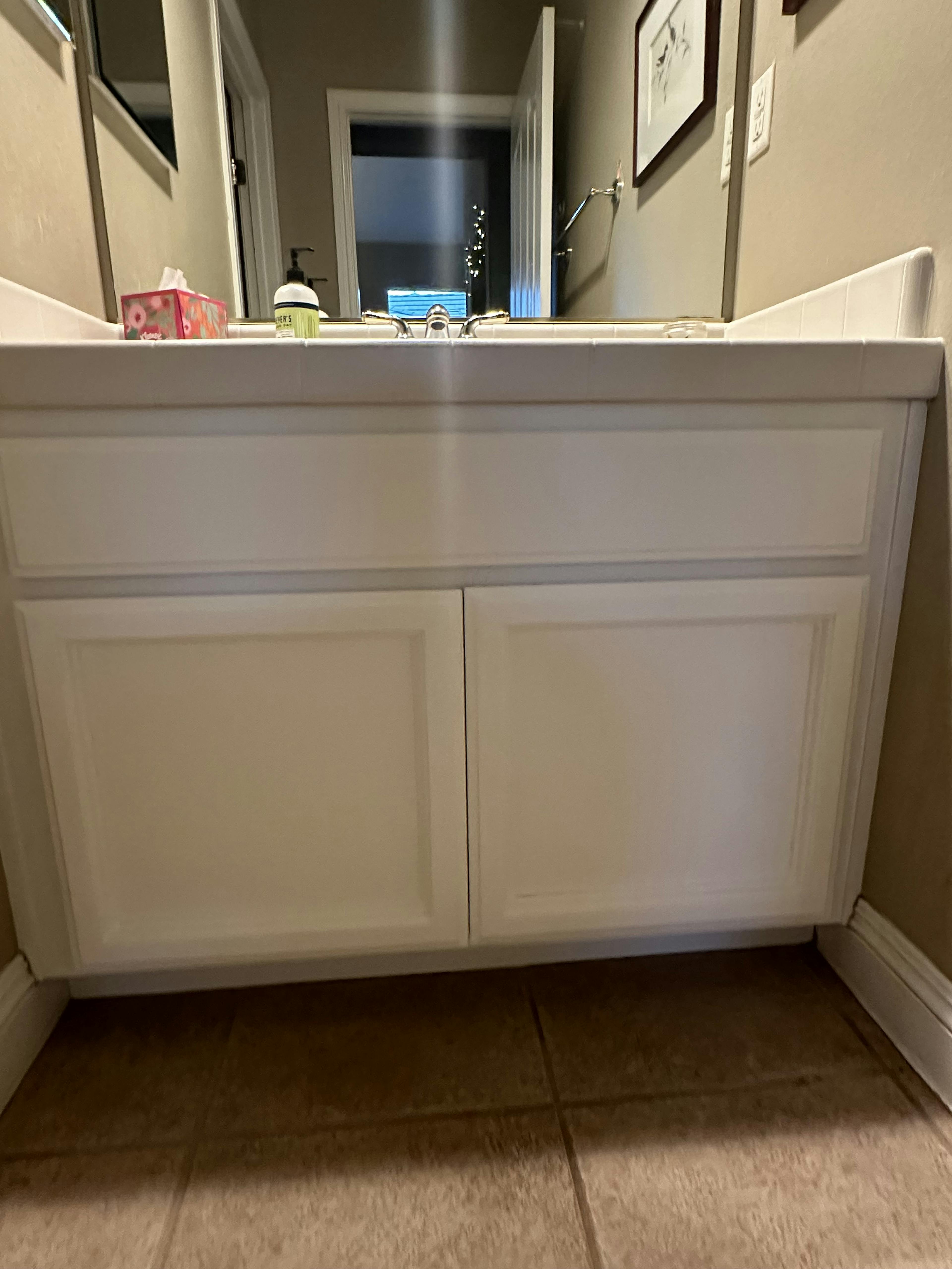 Bathroom vanity view with white cabinets, a countertop holding various toiletries, and a mirror reflecting a lit room. The floor is tiled, and the perspective is from a low angle, focusing on the cabinetry details.