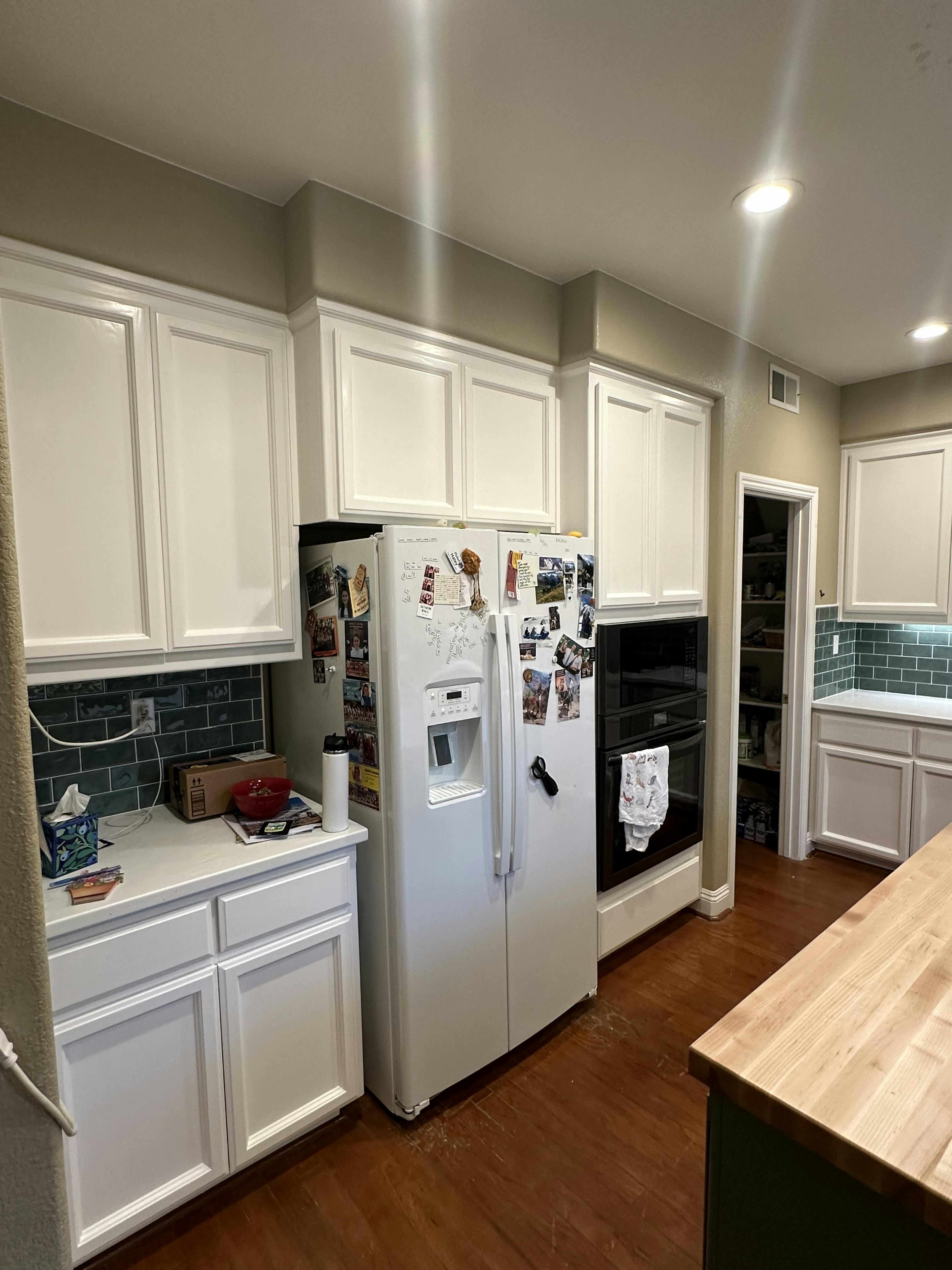 A well-lit kitchen corner with white paneled cabinets and a refrigerator covered in magnets and notes. The counter is cluttered with daily items, and a dark green subway tile backsplash is partially visible. The scene suggests a lived-in space with personal touches.