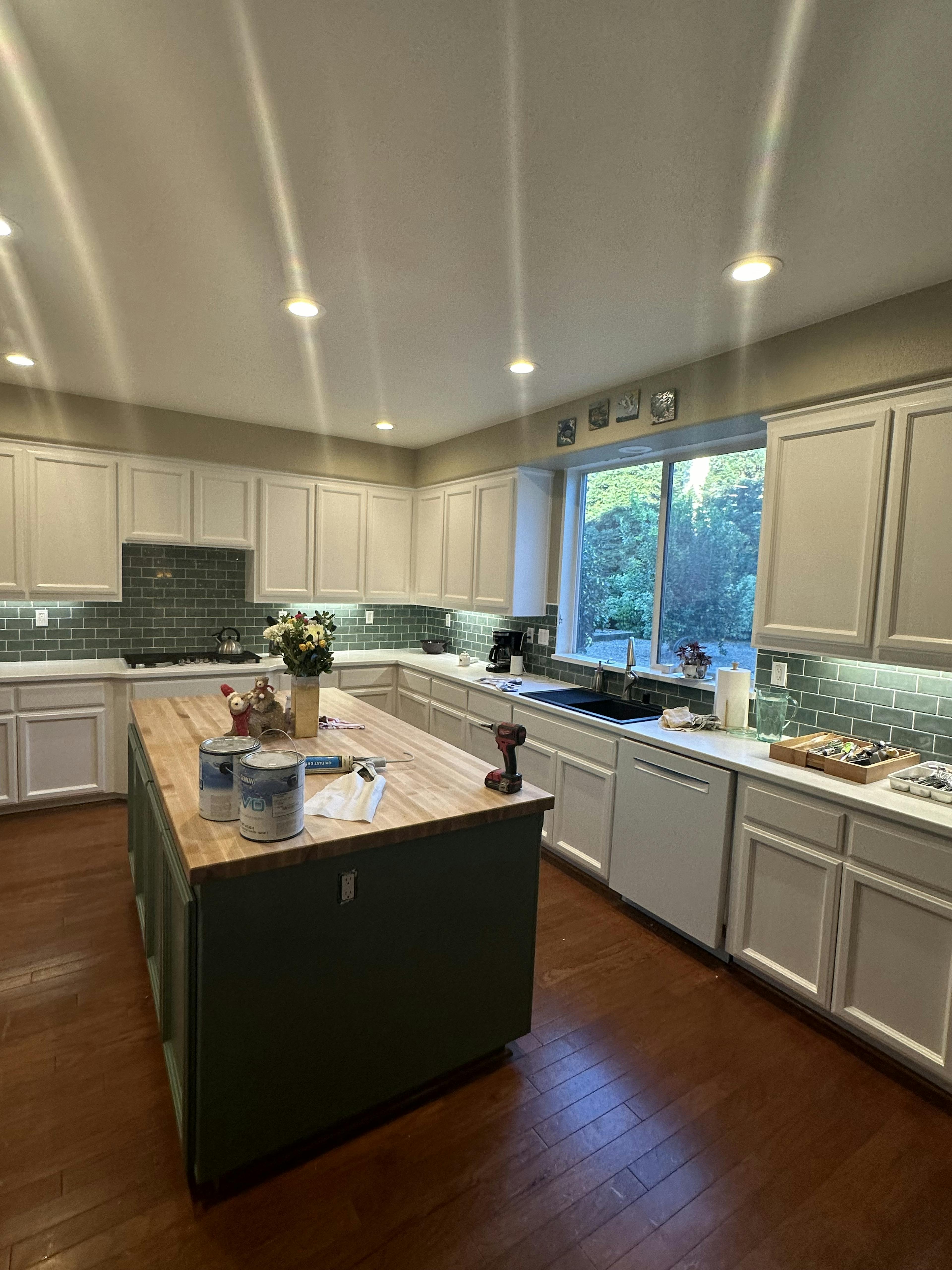 A kitchen with white cabinets and dark green backsplash under recessed lighting. The wooden island is the centerpiece, covered with painting supplies and a vase of flowers, indicating ongoing decoration or renovation. Sunlight streams in through a window with a view of greenery outside.