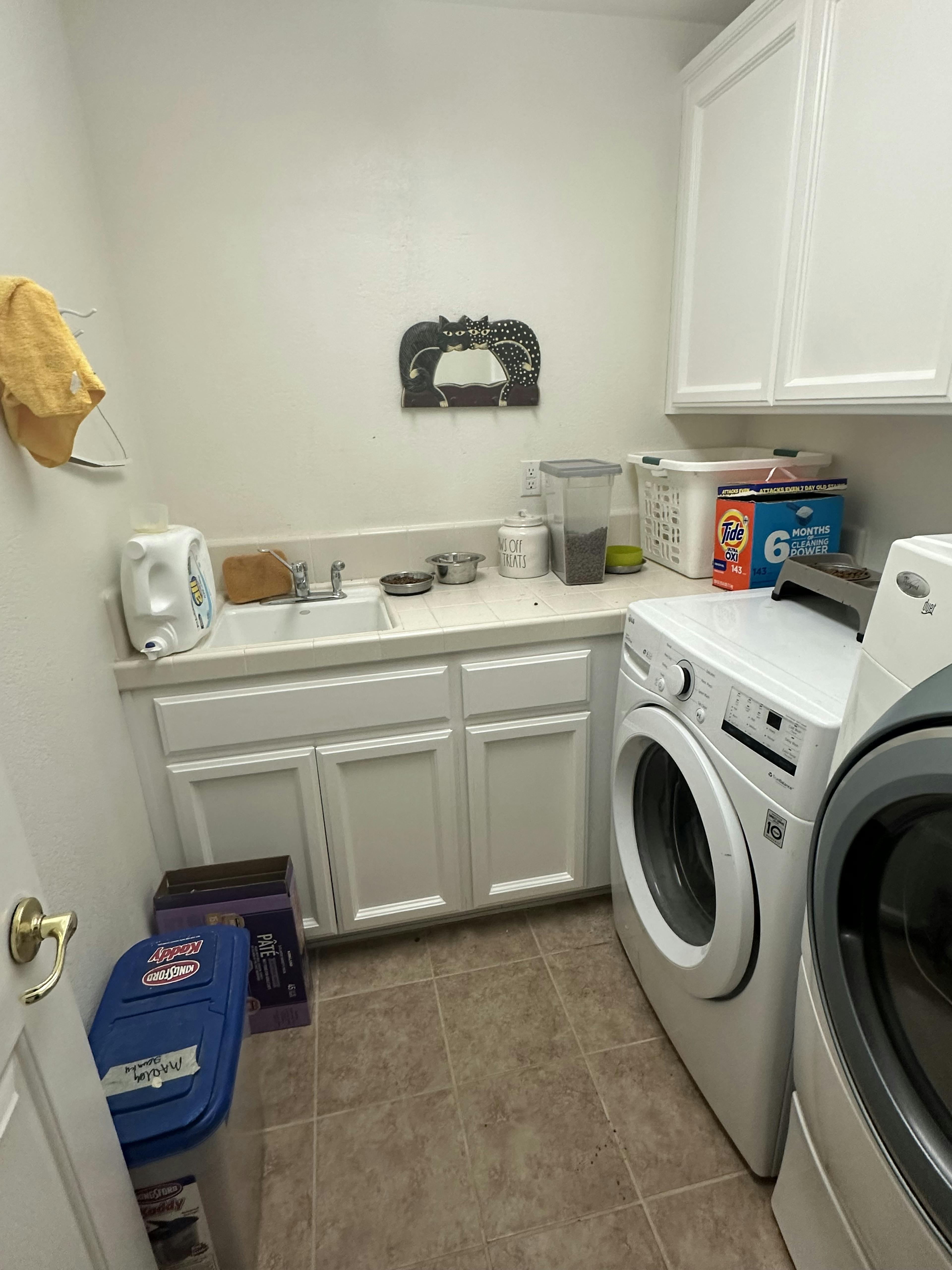 Functional laundry room with white cabinetry and a built-in sink, a washer, and a dryer. Essentials like detergent and a fabric softener are on top of the dryer. A whimsical cat-shaped wall decor adds a personal touch to the room.