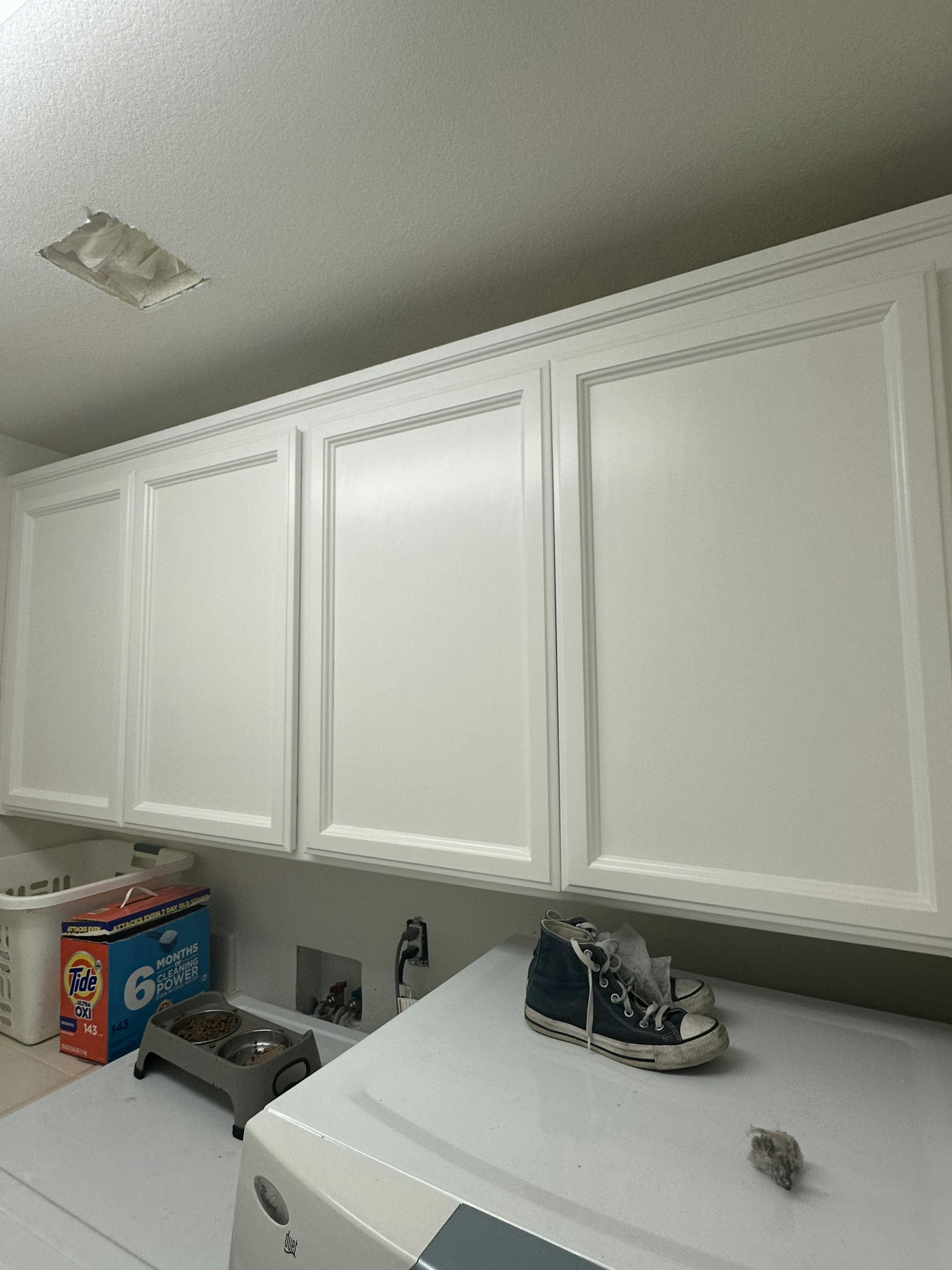 Laundry room with white cabinets above a gray appliance, presumably a washer or dryer. On the appliance, a box of detergent and a purple high-top sneaker are placed next to a small pile of lint, with a partially visible utility sink and wall outlet to the left.