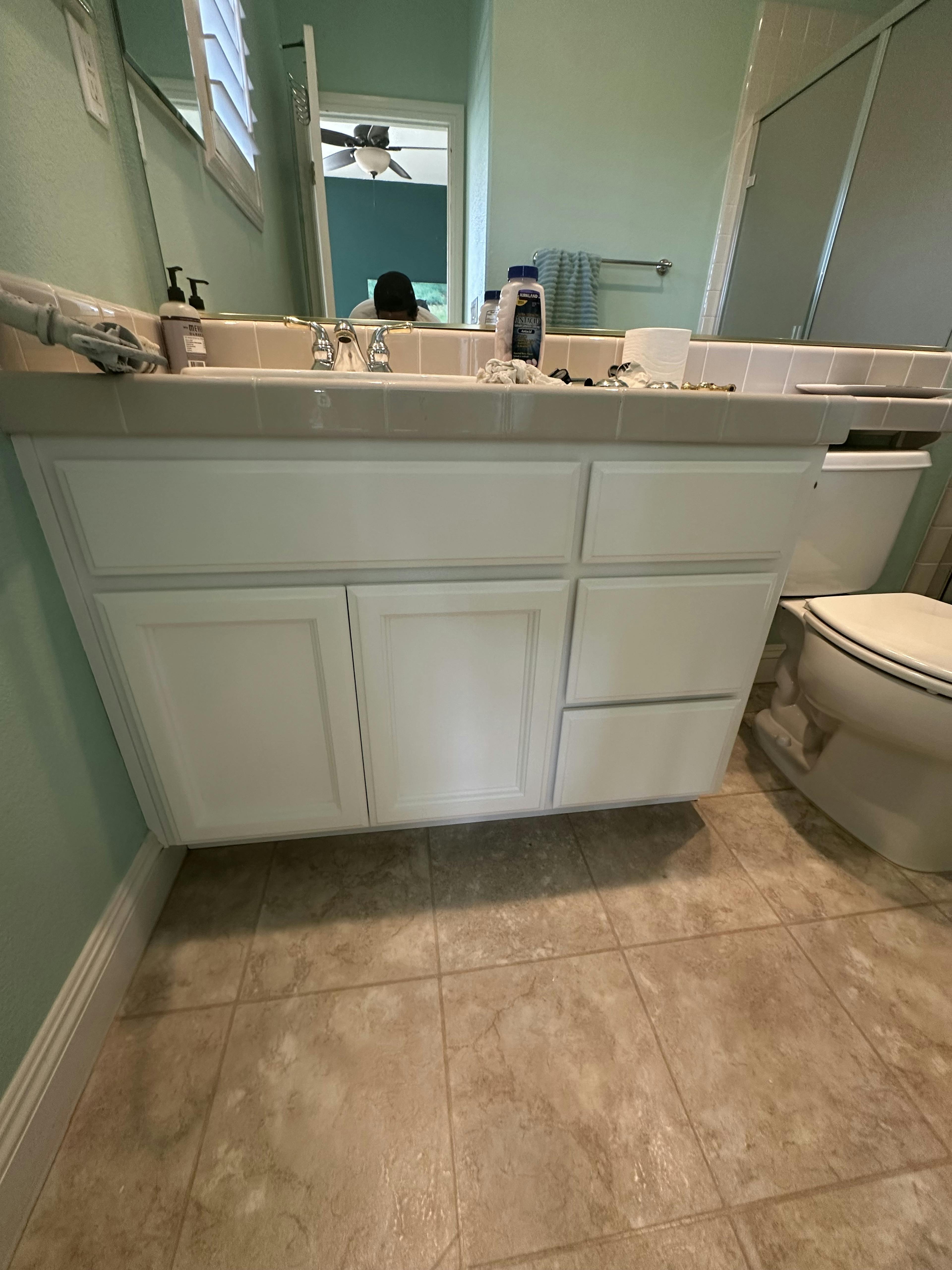 Bathroom interior with a mint green wall and a large vanity featuring white cabinetry. Above the vanity is a mirror reflecting a ceiling fan, with various toiletries and a hand towel on the countertop. To the right, there's a white toilet next to a tiled floor.