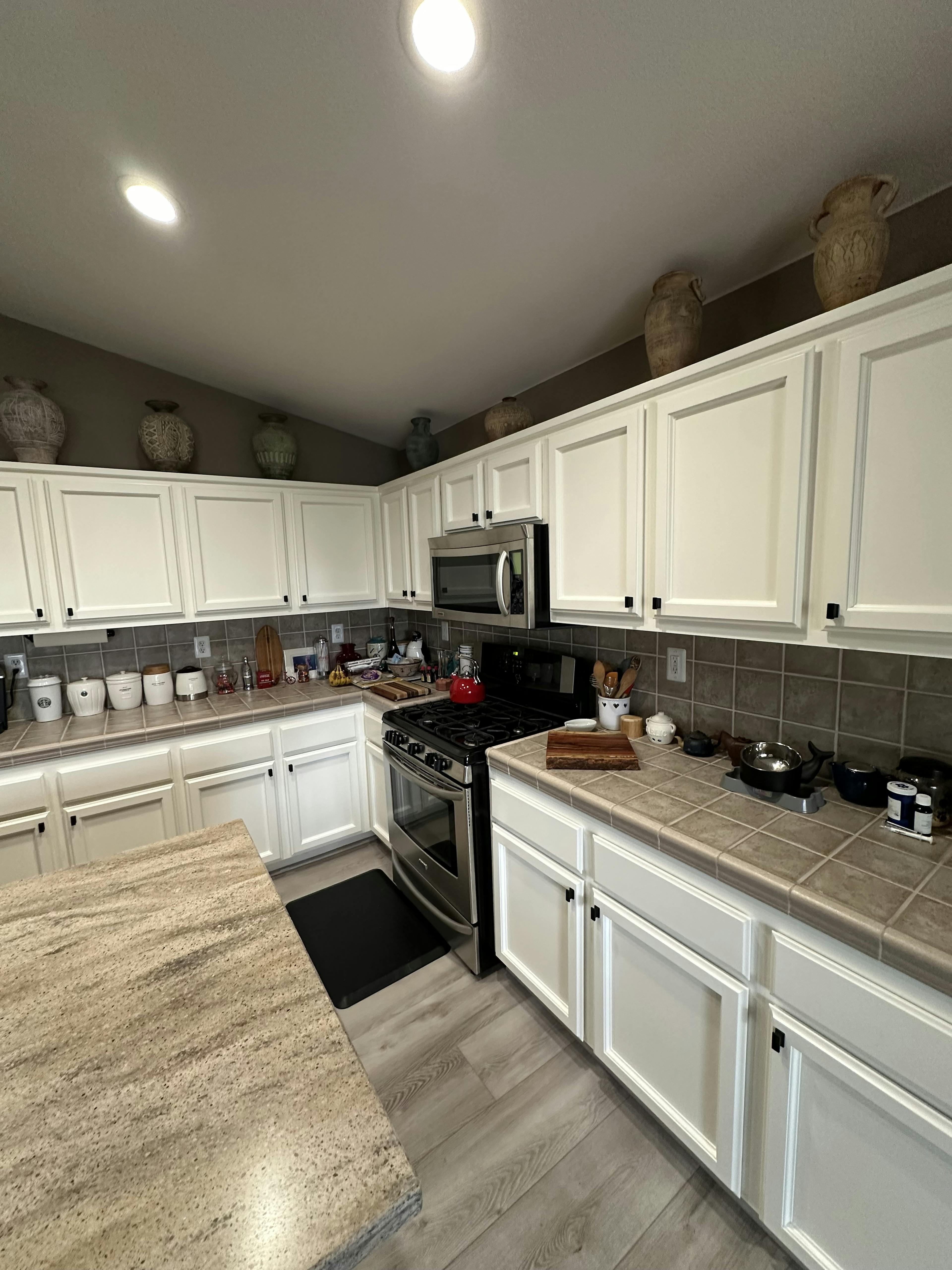 A kitchen with white cabinetry, beige countertops, and gray floor tiles. A gas stove, microwave, and various kitchen utensils are visible. Decorative vases sit above the cabinets, adding a personal touch to the space, which is brightly lit by recessed ceiling lights.