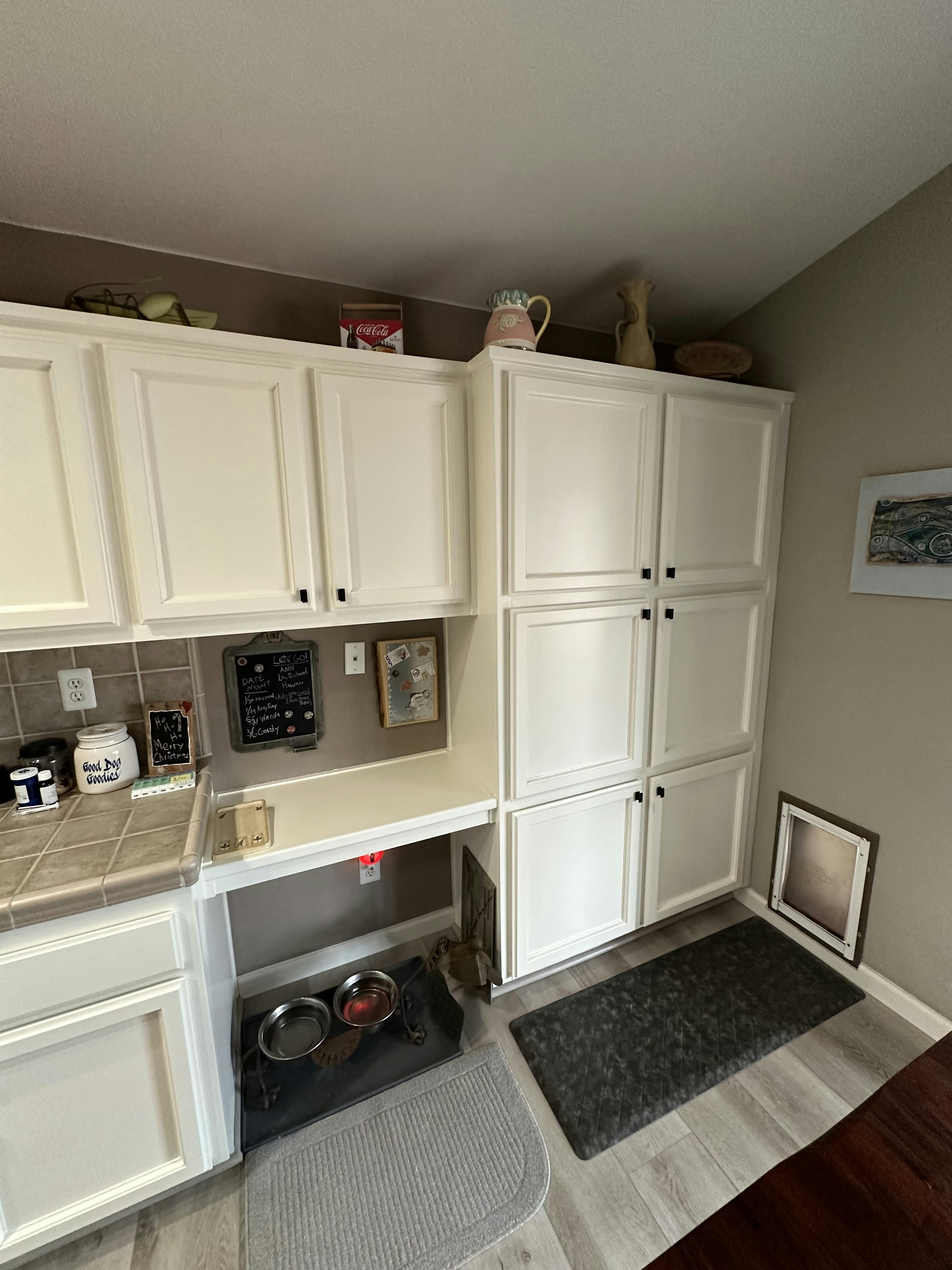 Corner of a kitchen with white cabinets, beige tile backsplash, and gray floor. There's a pet feeding station with bowls on a mat, a small chalkboard, and decorative items on the countertop. Above, eclectic objects decorate the top of the cabinets.