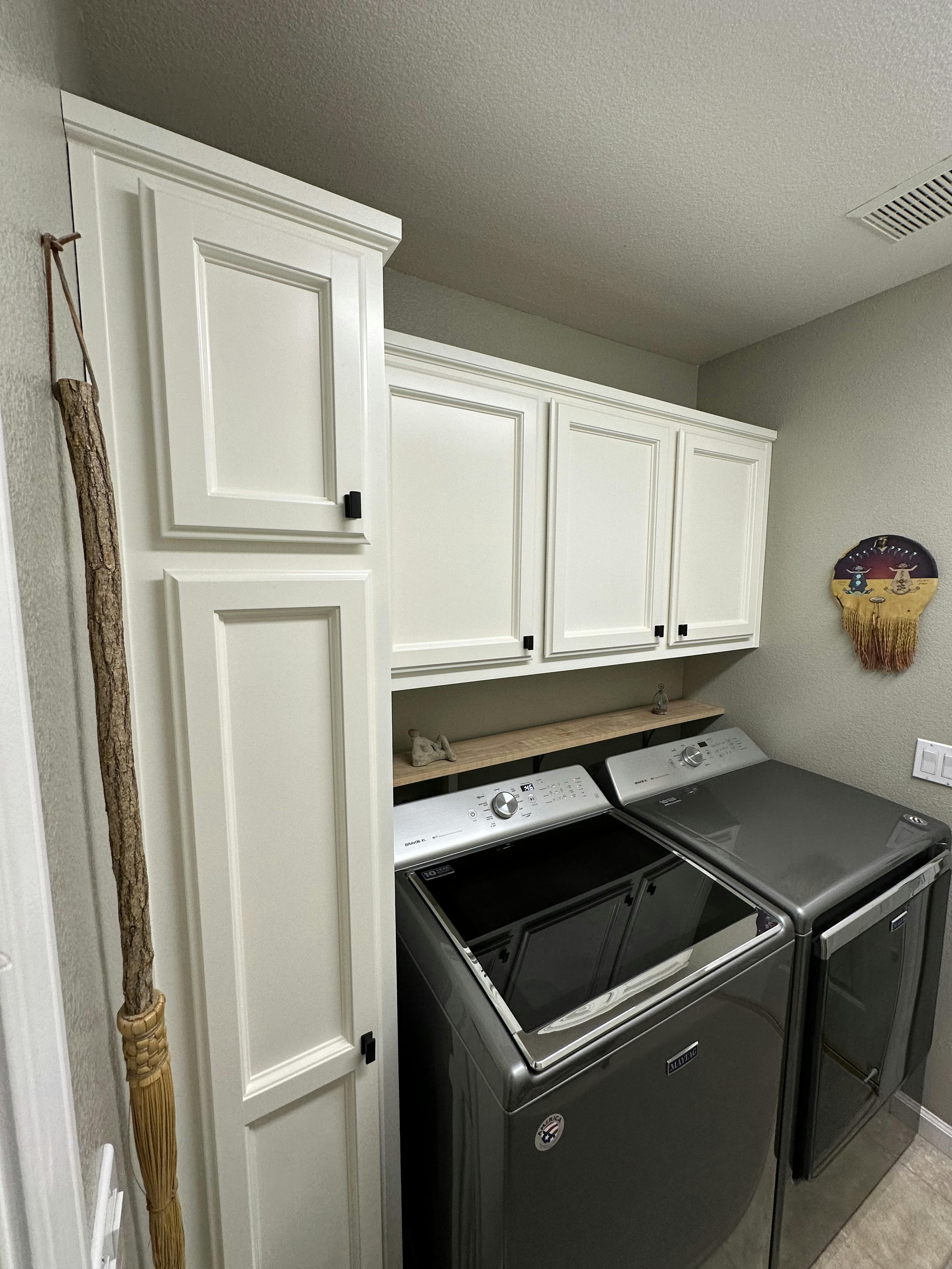 Laundry room with white cabinets above a set of dark grey washer and dryer. A wooden broom leans against the wall on the left, and a whimsical wall art piece is visible on the right. The appliances are modern and top-loading, set against a light grey wall.