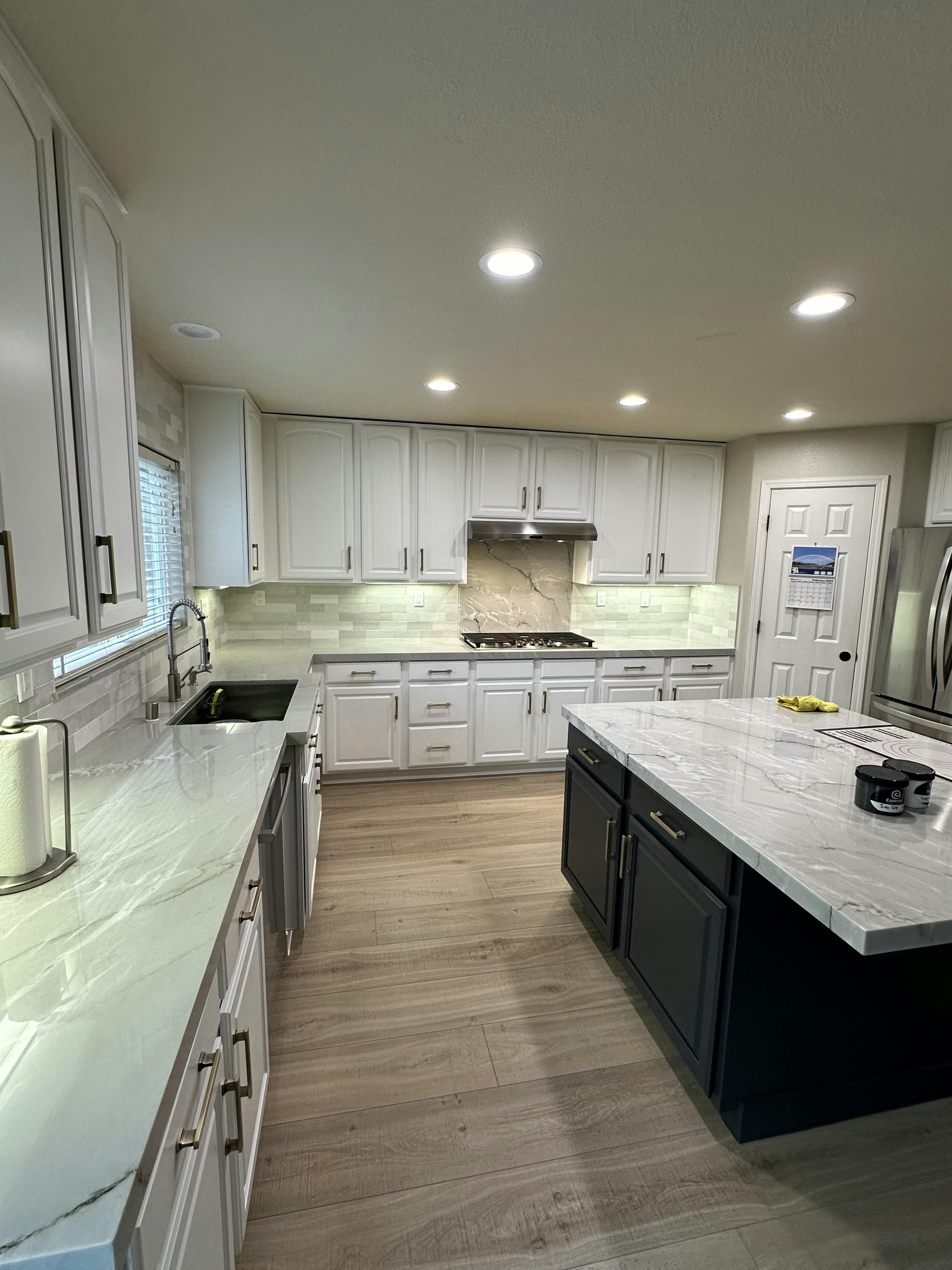 Elegant kitchen with white upper cabinets and a contrasting dark island, featuring marble countertops and wood flooring. Recessed lighting brightens the space.