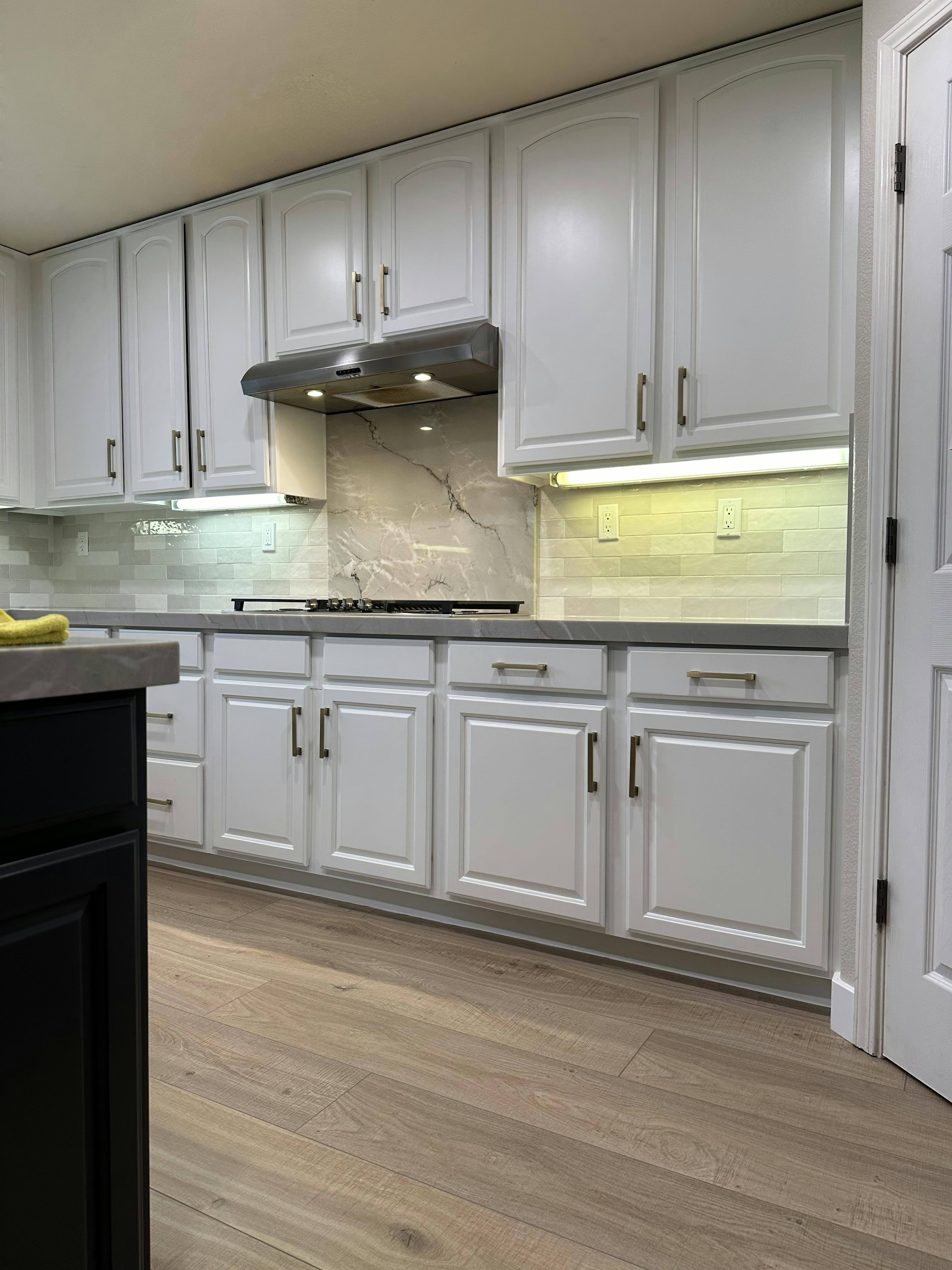 Kitchen stove area with white cabinetry, under-cabinet lighting, and a stainless-steel range hood against a light subway tile backsplash. The marble-like backsplash behind the stove adds a luxurious touch.