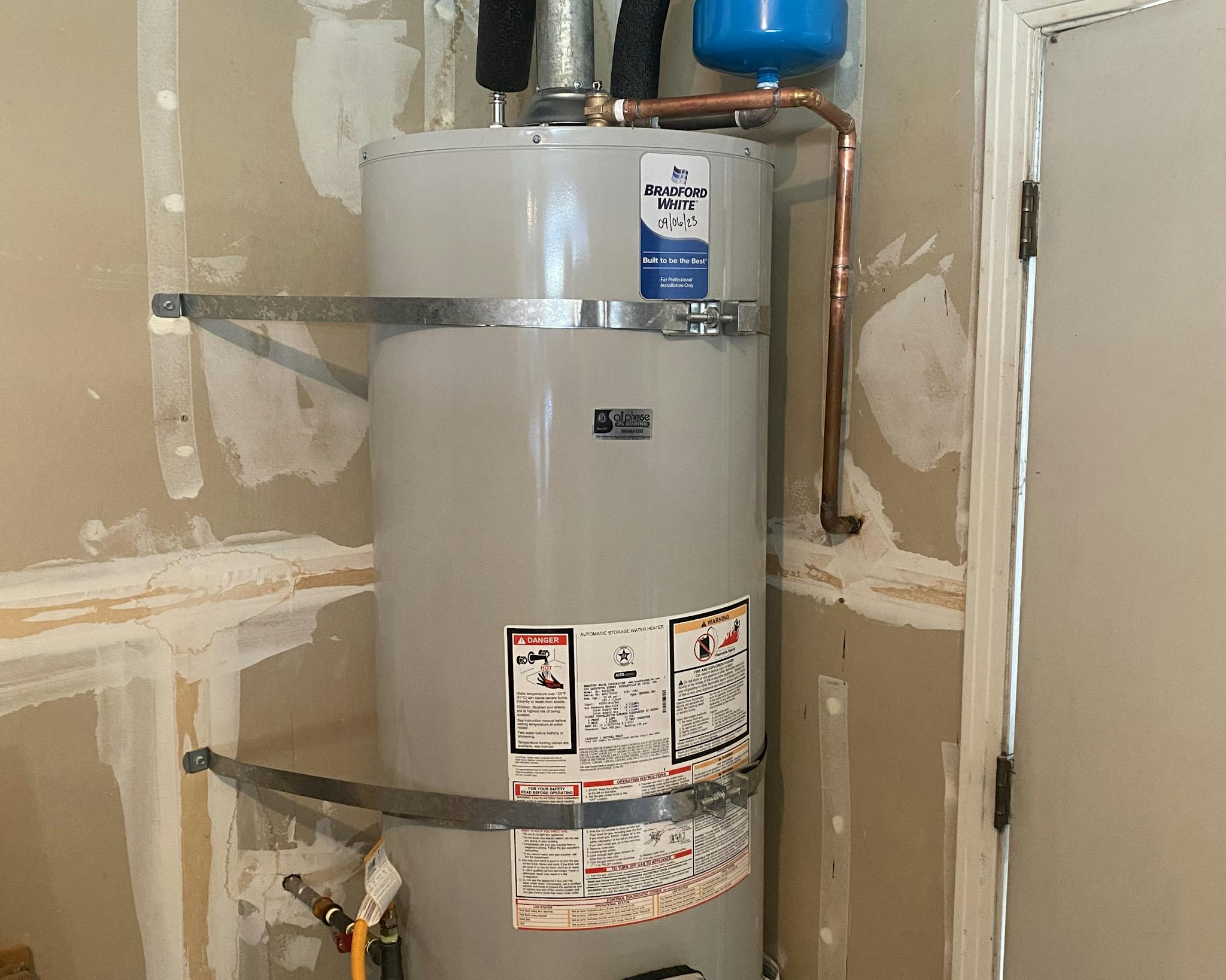 A large cylindrical 'Bradford White' water heater mounted against a wall with unfinished patchwork. The heater has a gray exterior with labels and safety instructions printed on it. Copper piping is visible at the top, and a small blue expansion tank is attached. A yellow valve handle can be seen near the bottom.