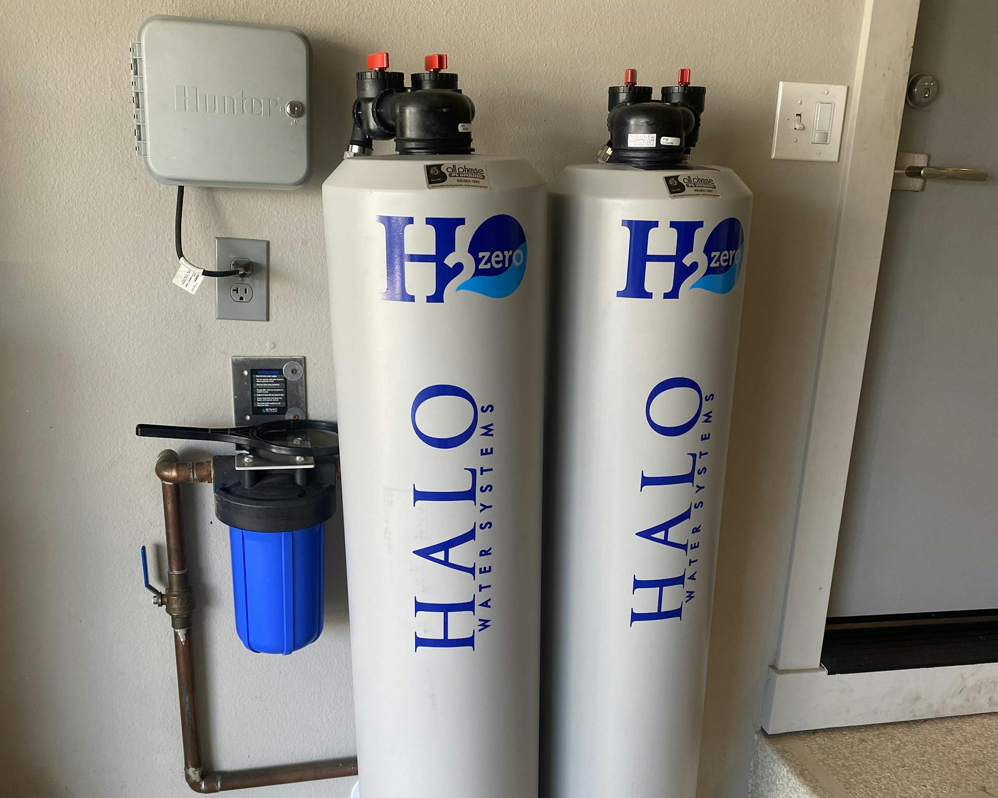 ""Two tall white cylinders labeled 'H2Ozero' and 'HALO Water Systems', placed side by side against a wall. Each cylinder has black caps with red valves. To the left, there's a smaller blue filter system connected to copper pipes and a control panel. Above the system, there's a gray electrical box on the wall.