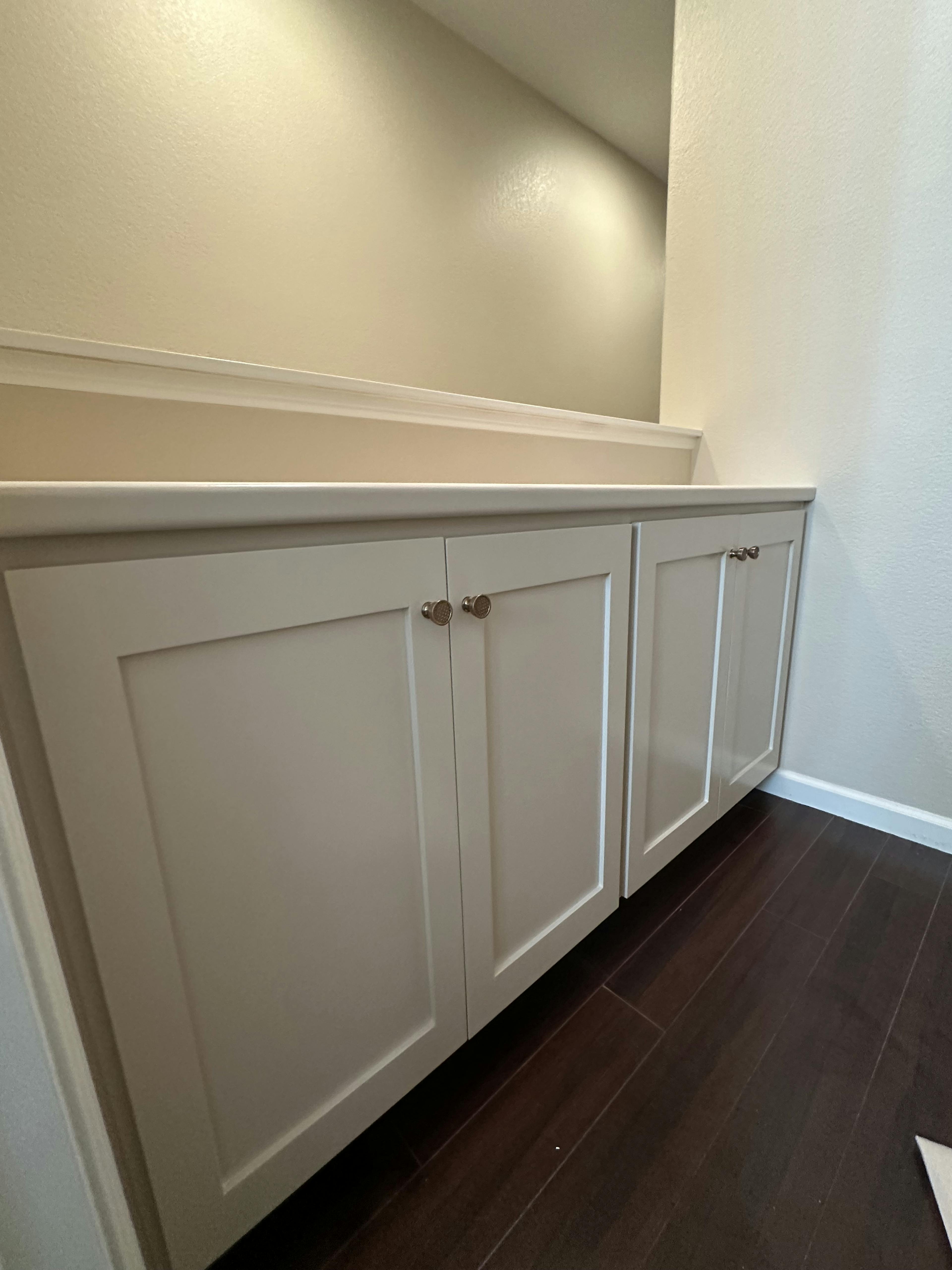 A narrow cabinet with white paneled doors and vintage handles, set against a warm beige wall and dark hardwood flooring, offering a simplistic and clean design.