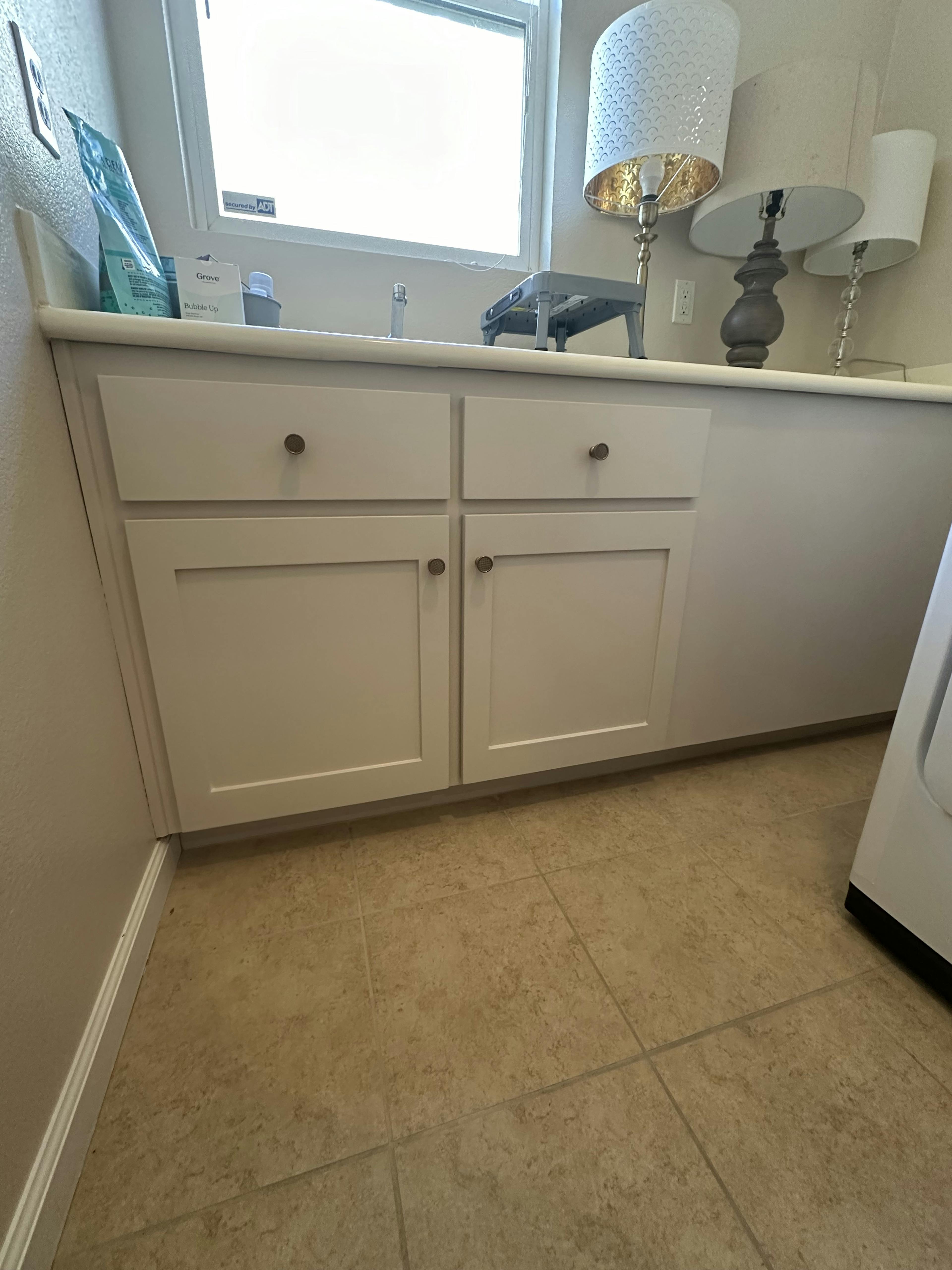 A well-lit, tidy laundry room with white cabinetry, a large window letting in natural light, and various cleaning supplies on the counter.