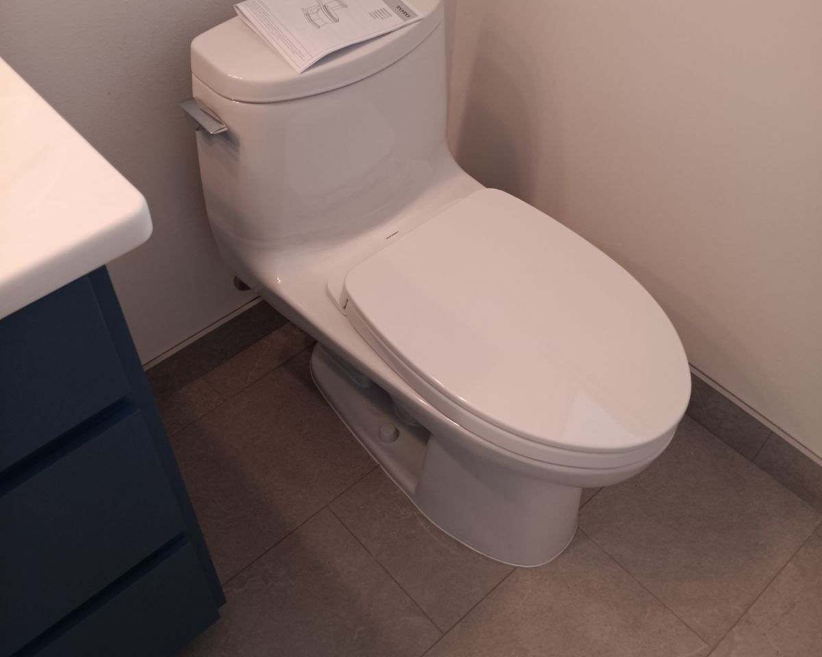 A modern, minimalist bathroom with a white toilet against a neutral wall. A manual rests on the closed lid, hinting at recent installation or maintenance. To the left, a glimpse of a grey vanity suggests a color-coordinated decor theme. The light brown tiled floor adds warmth to the space.