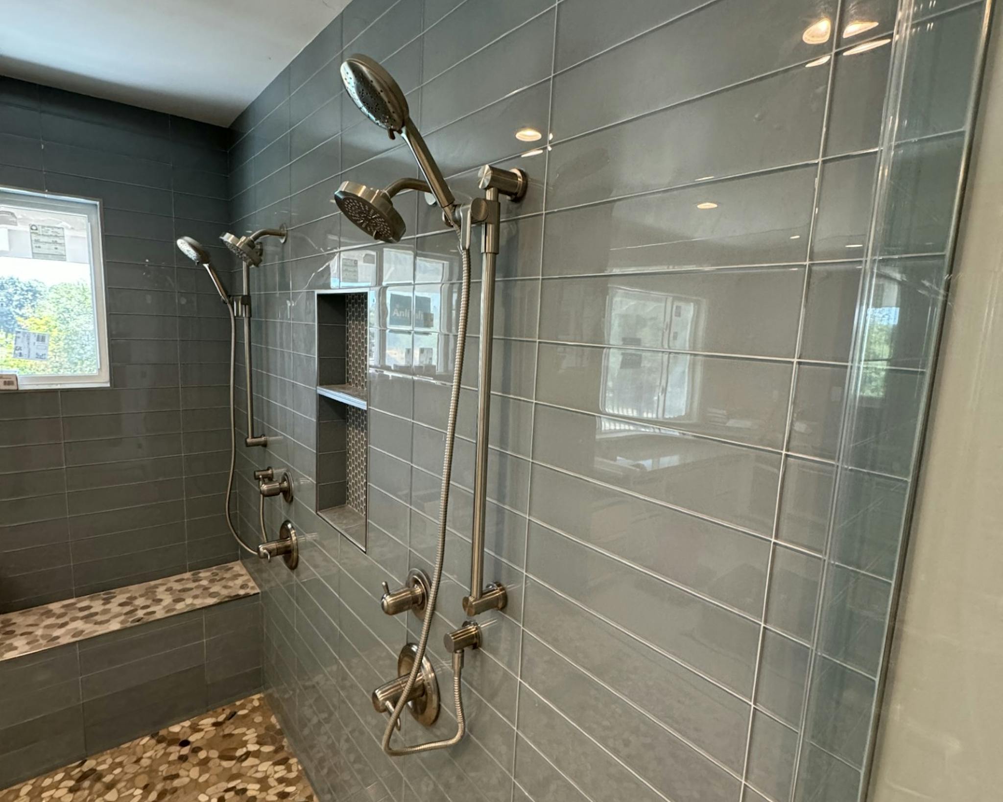 Modern, spacious bathroom featuring a rectangular bathtub with multiple wall-mounted shower heads, safety grab bars, and a mosaic stone floor. The gray tile walls and large windows provide a clean, contemporary feel.