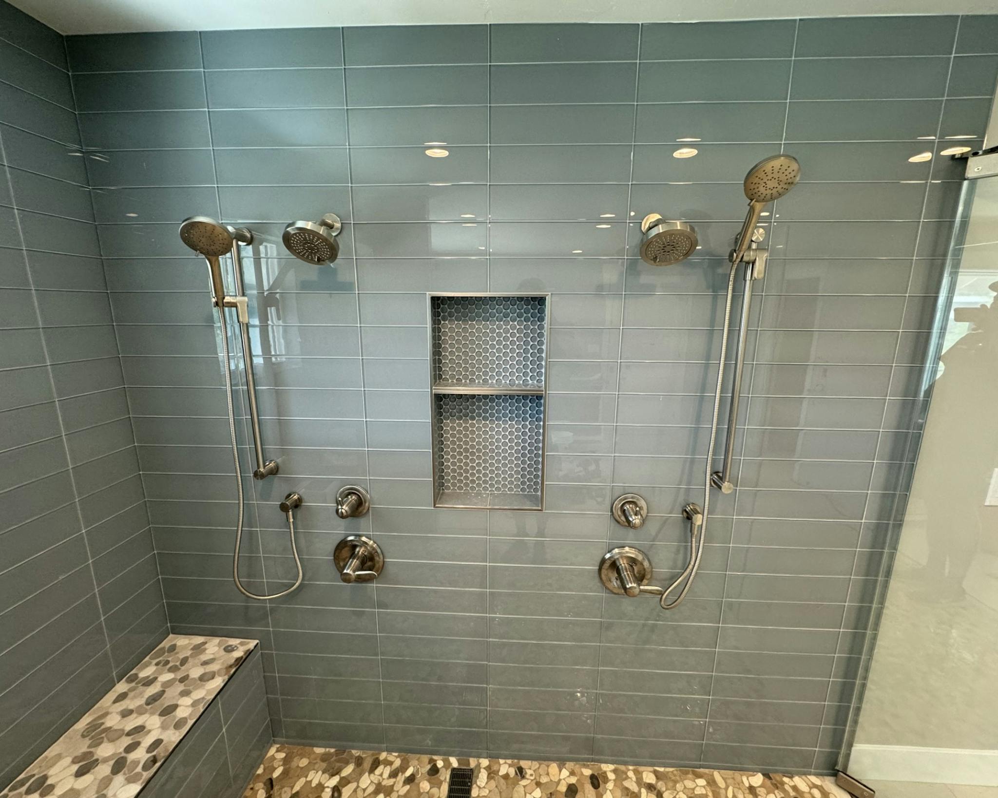 View of a modern shower setup in a bathroom, showcasing three adjustable shower heads mounted on a sleek, gray-tiled wall with built-in shelves for toiletries. The design includes safety grab bars and a built-in bench, reflecting a functional yet stylish space."