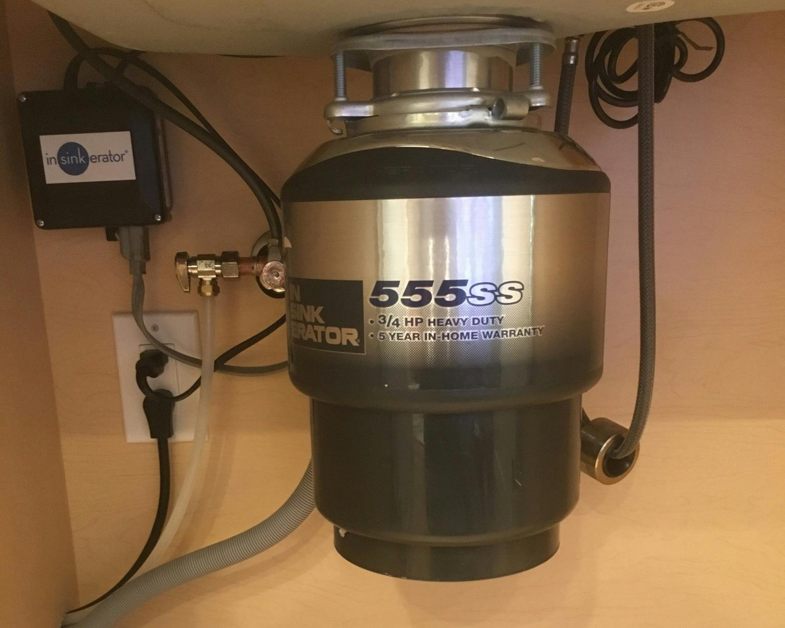 A close-up view of an 'InSinkErator' garbage disposal unit, model 555ss, boasting 3/4 HP heavy-duty performance with a 5-year in-home warranty label. The unit is mounted under a sink with a black power box labeled 'insinkerator' to its left, and various cords and tubes connected.