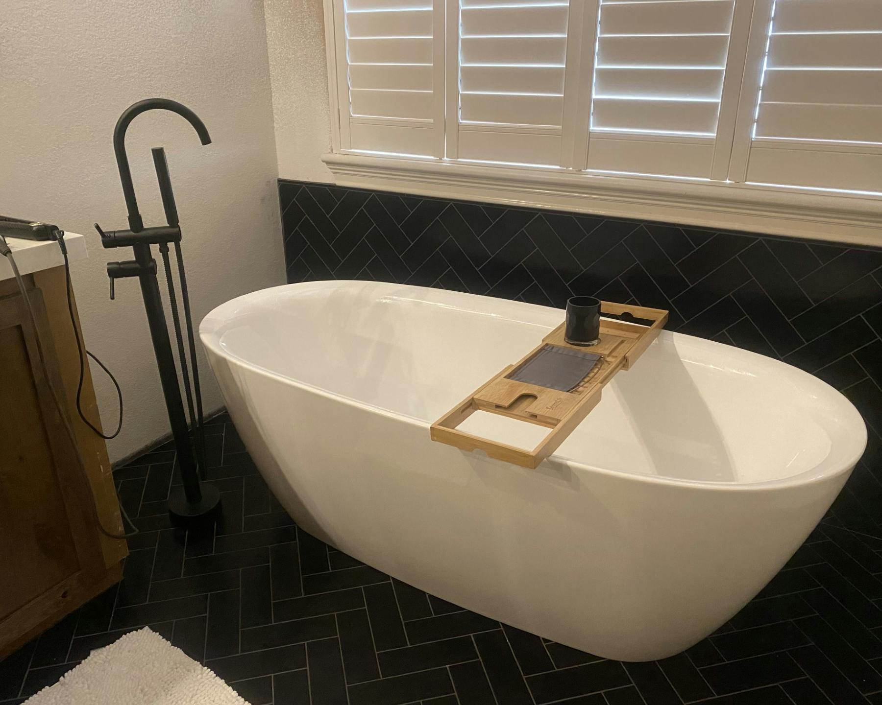 A modern bathroom featuring a sleek white freestanding bathtub against a wall with white shutters. A wooden bath caddy rests atop the tub, holding a dark-colored mug. To the left of the tub stands a tall, dark-colored floor-mounted faucet. The room has black tiles with a herringbone pattern accent, and a fluffy white bath mat lies on the floor.