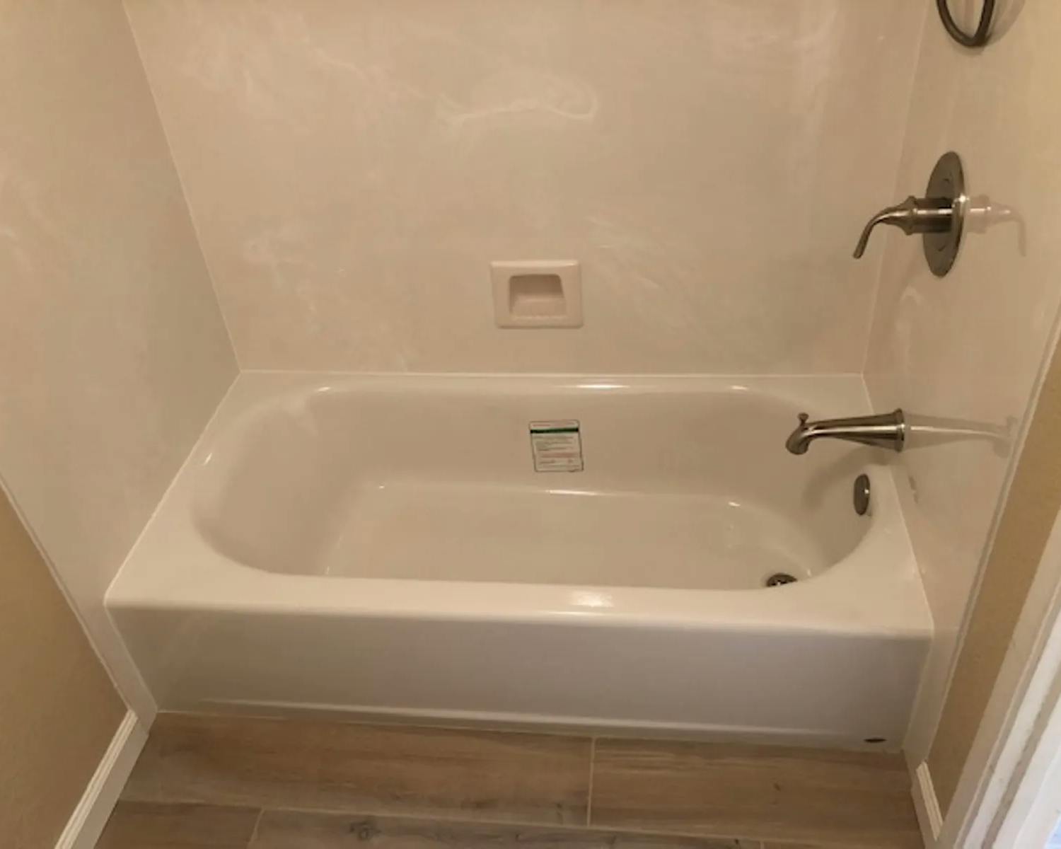 A white bathtub with a built-in shower, set against a beige wall. A small recessed shelf is on the wall for holding items, and a label is attached inside the tub. Bronze-colored fixtures, including the faucet and showerhead, are installed on the right side of the tub.