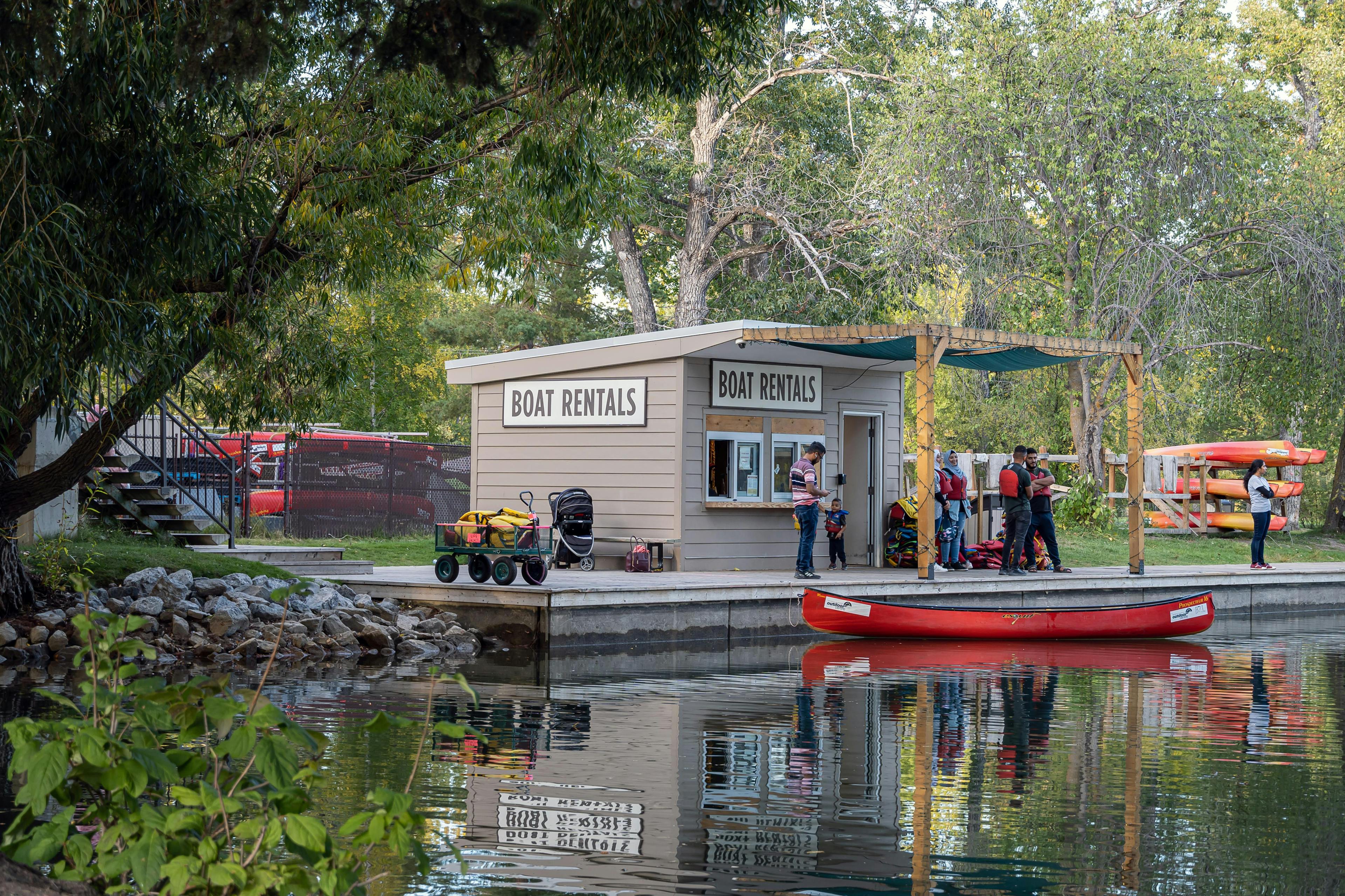 A group of people in life jackets queue up at a boat rental dock, beneath dense green trees, alongside a river, near a red canoe.