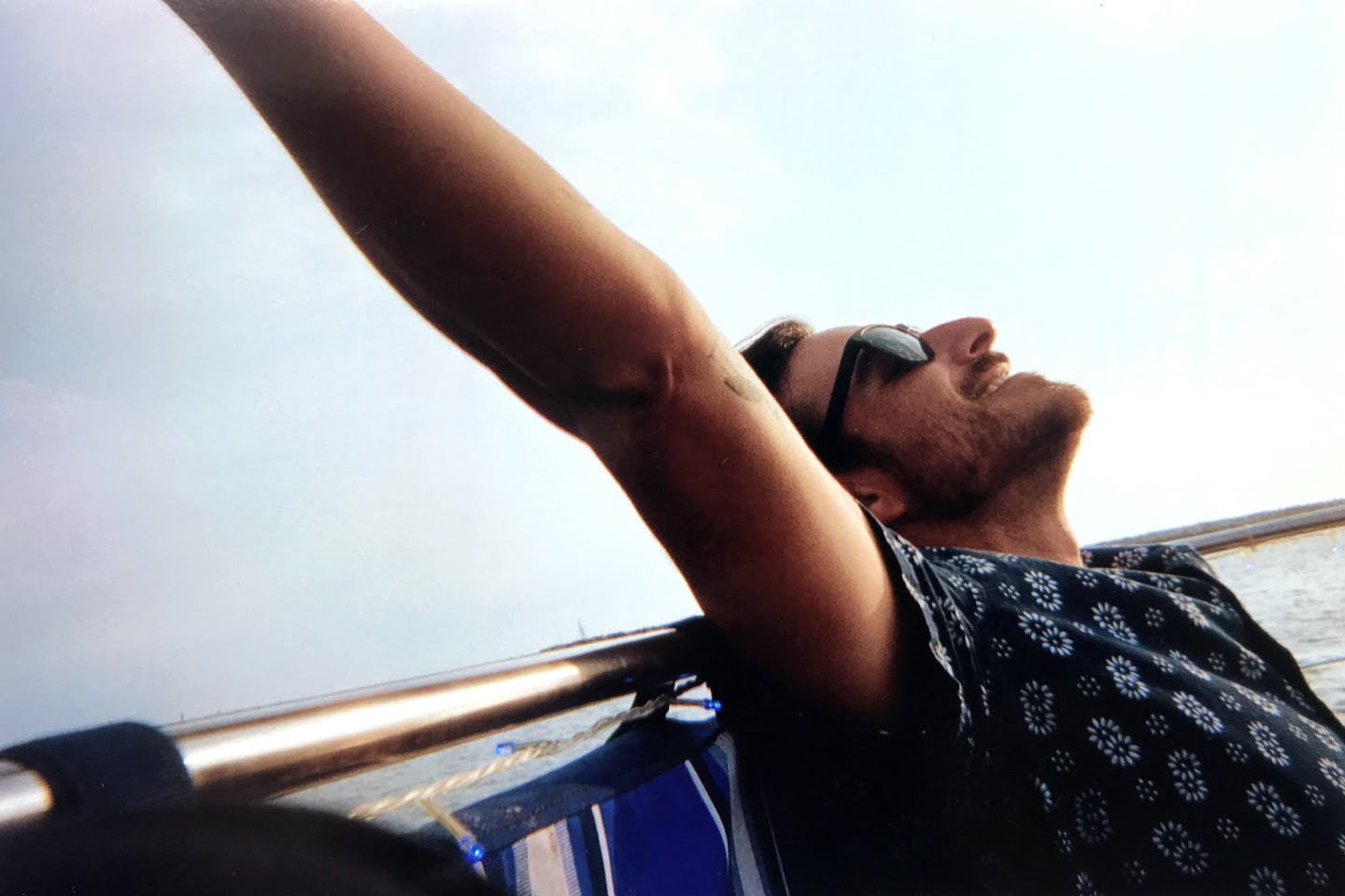 A man in sunglasses is smiling with his arms in the air. He is on a boat. He is wearing a navy shirt with white flowers on it.