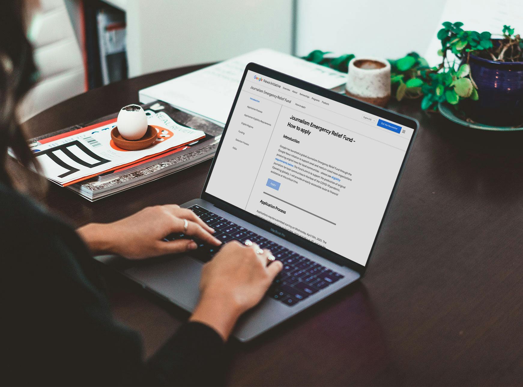 A woman types on a laptop, with the Journalism Relief Emergency Fund in the laptop. Behind the laptop is a plant, a few magazines with a coffee cup on top, and a candle. The woman is wearing a black shirt and has rings on her fingers.