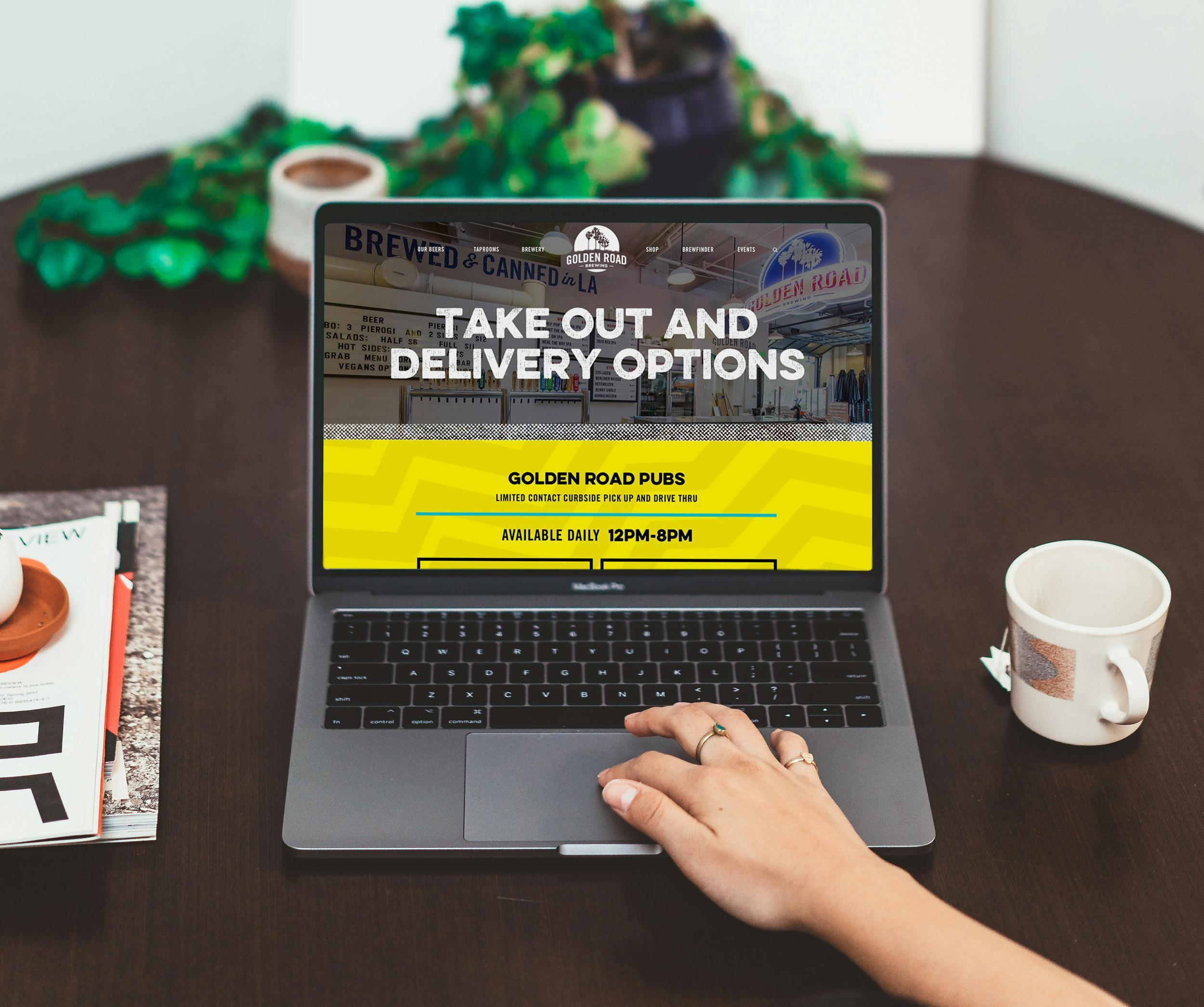 A hand is on the scroll pad of a laptop with the Golden Road homepage. There is a coffee mug to the right of the laptop. A plant in the background.