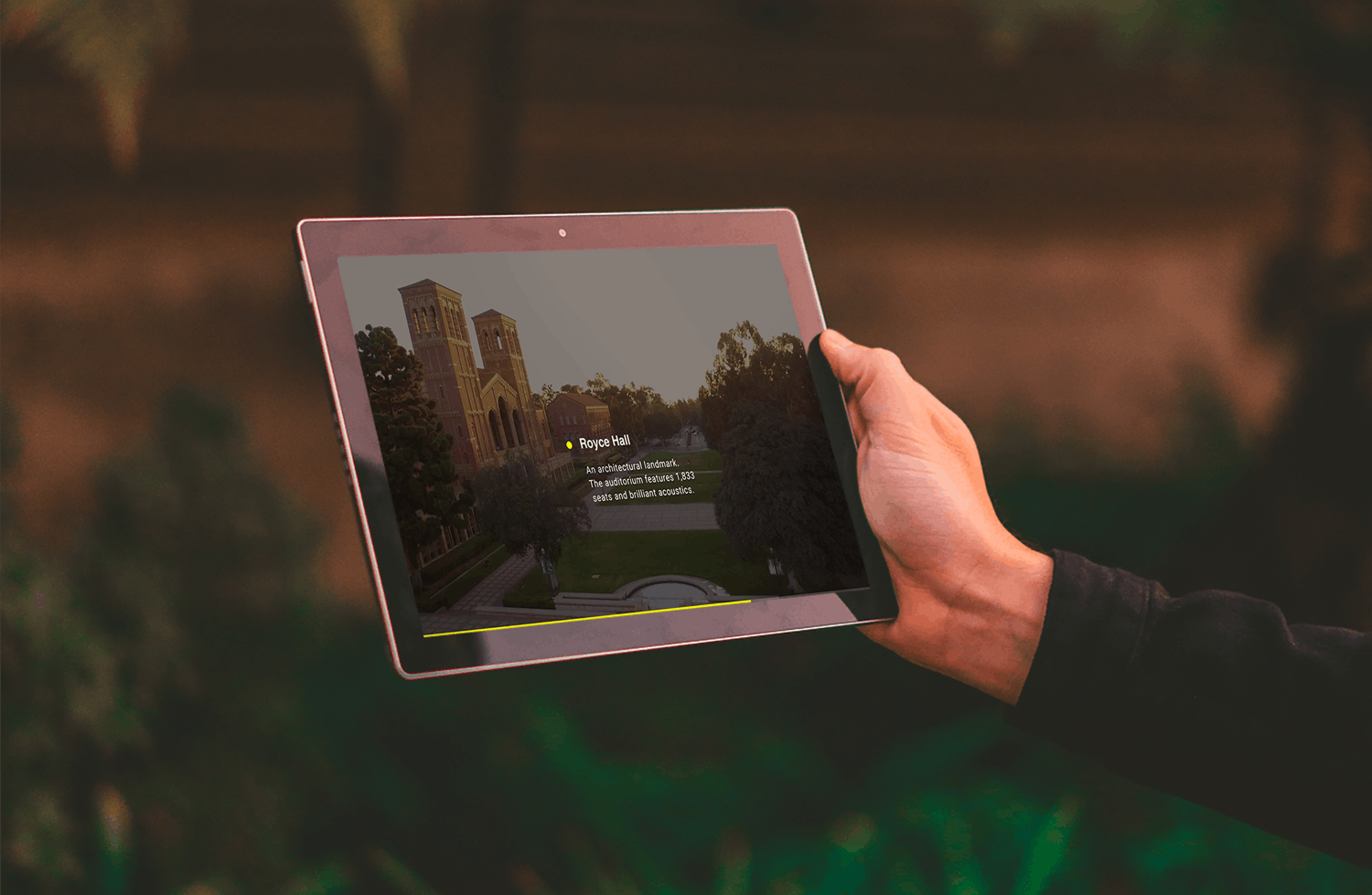 A hand holds an iPad with grass blurred out behind the iPad. The UCLA campus is shown on the iPad.