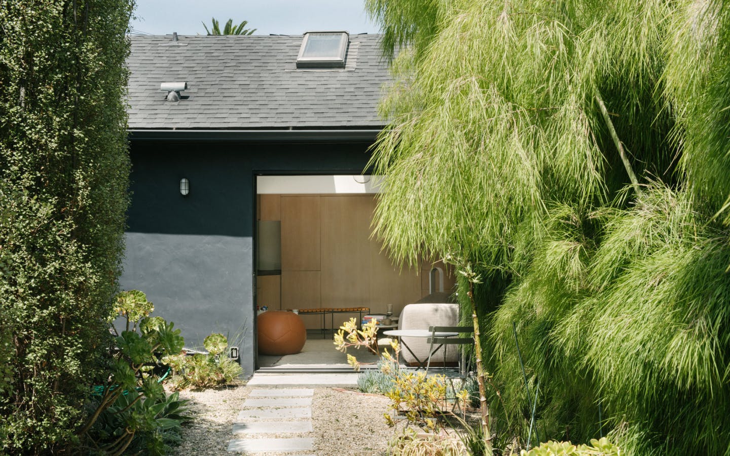The outside of a bungalow in Venice Beach. There are trees in the foreground and a black bungalow with a large garage door open. Inside you can see two stools and a leather beanbag chair. In the foreground there's also a little bistro table set