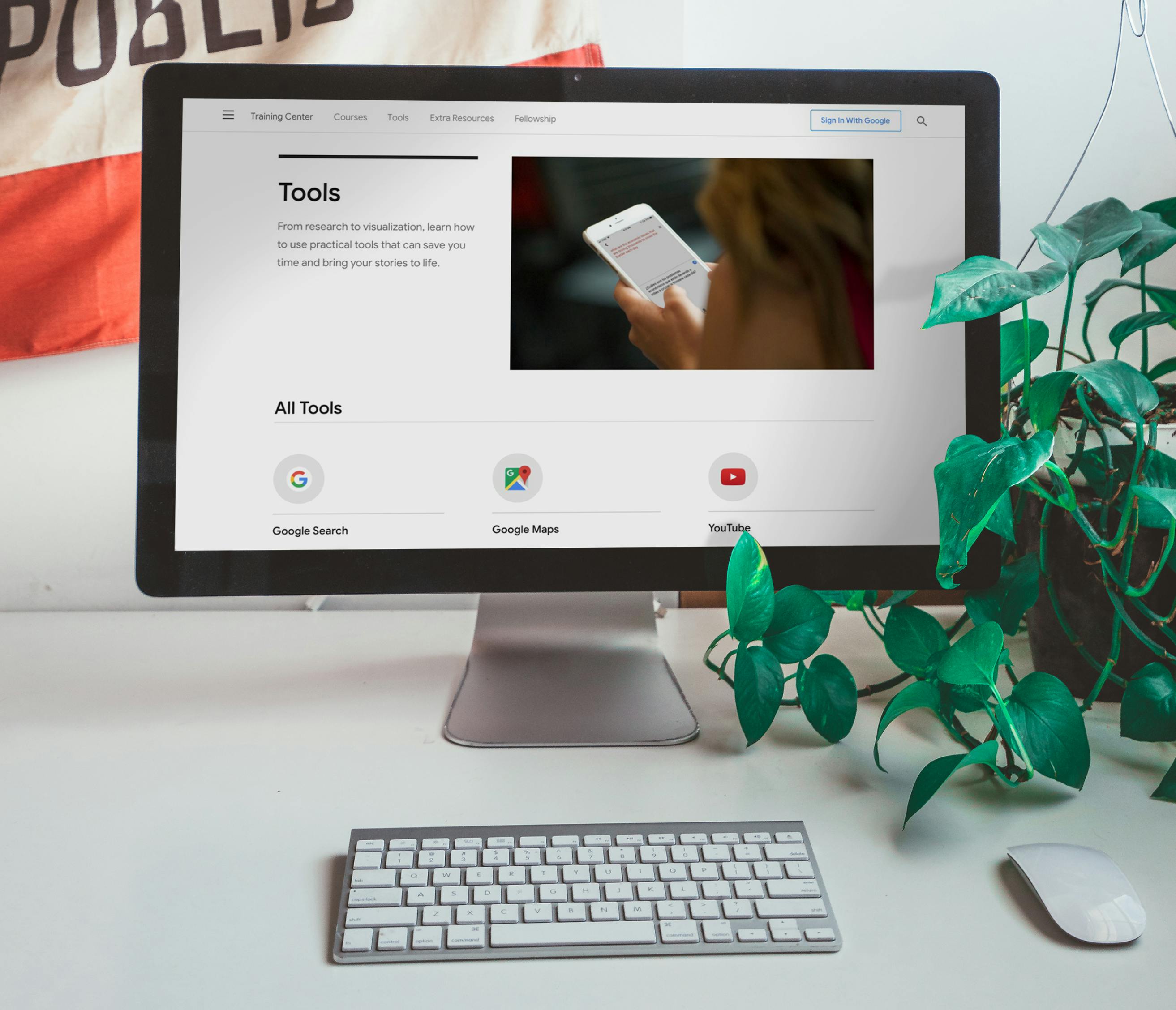 A monitor is on a white desk with a plant to the right, a keyboard and mouse in front, and a corner of the California flag behind it. On the monitor is a website with a list of Google Tools.