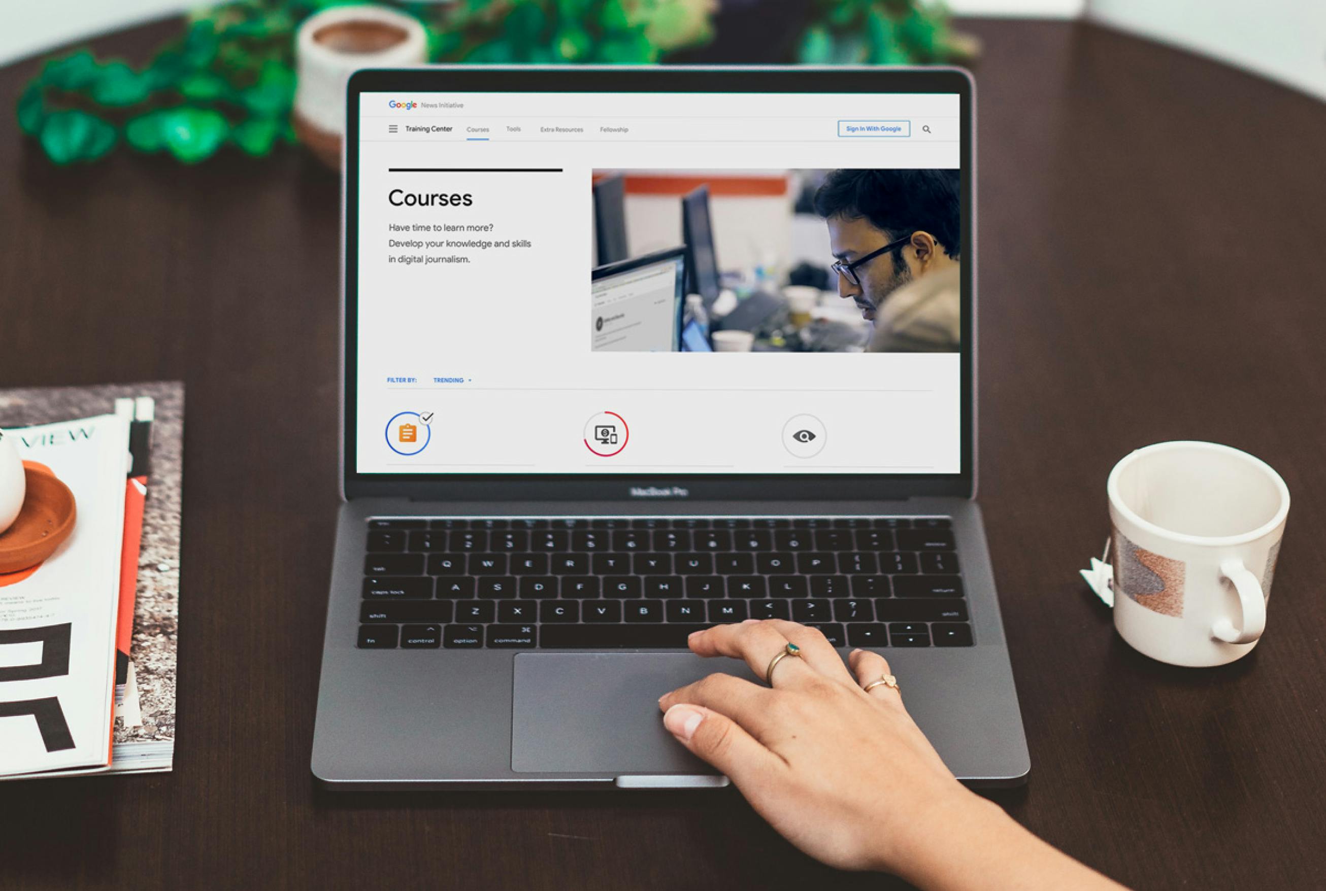 A hand is on a trackpad of a laptop. To the right of the laptop is a coffee mug, to the left is a stack of magazines. On the laptop is a website with Courses and icons.
