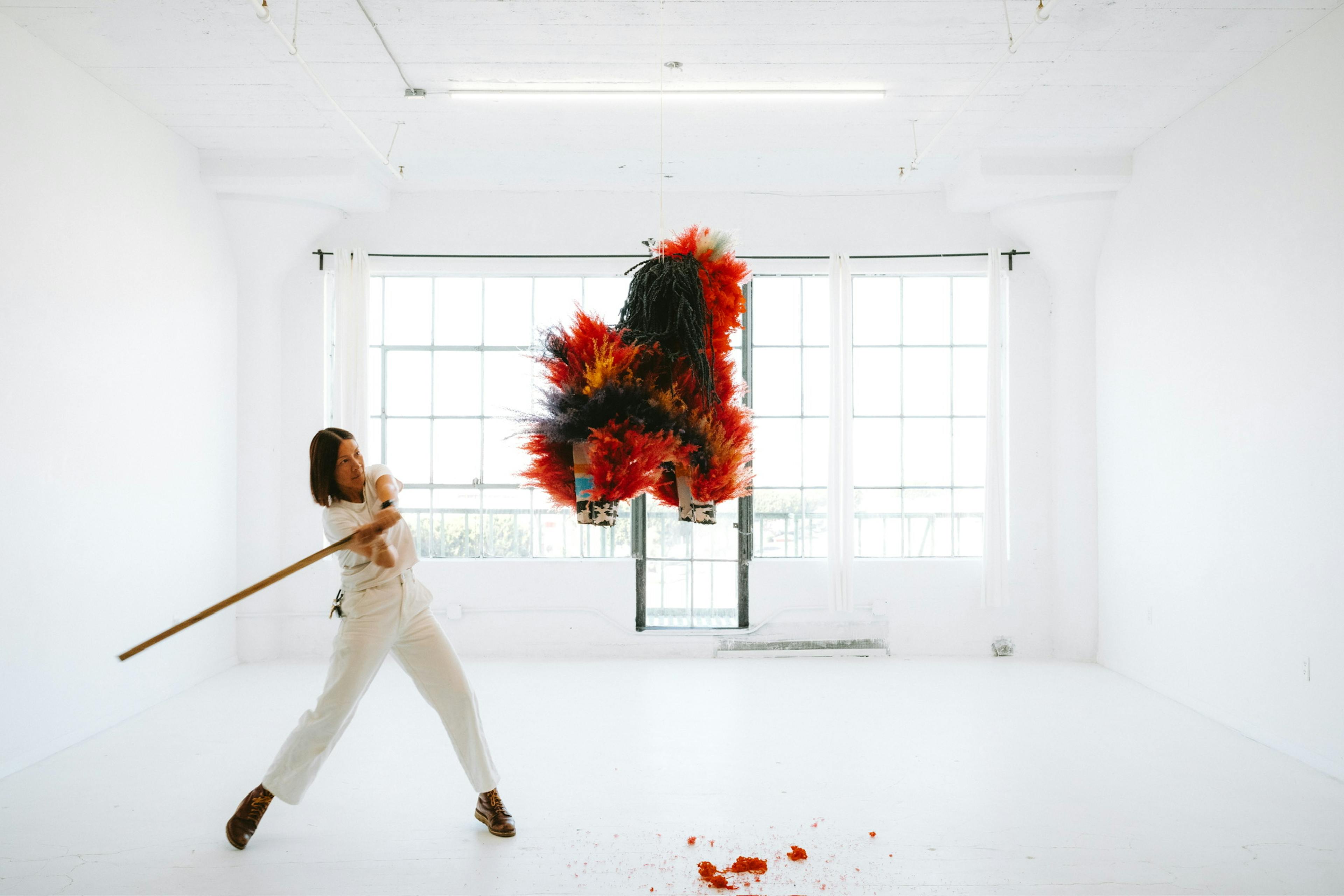 A women hitting a bright red flower-piñata with a piñata stick in a white room with a window.