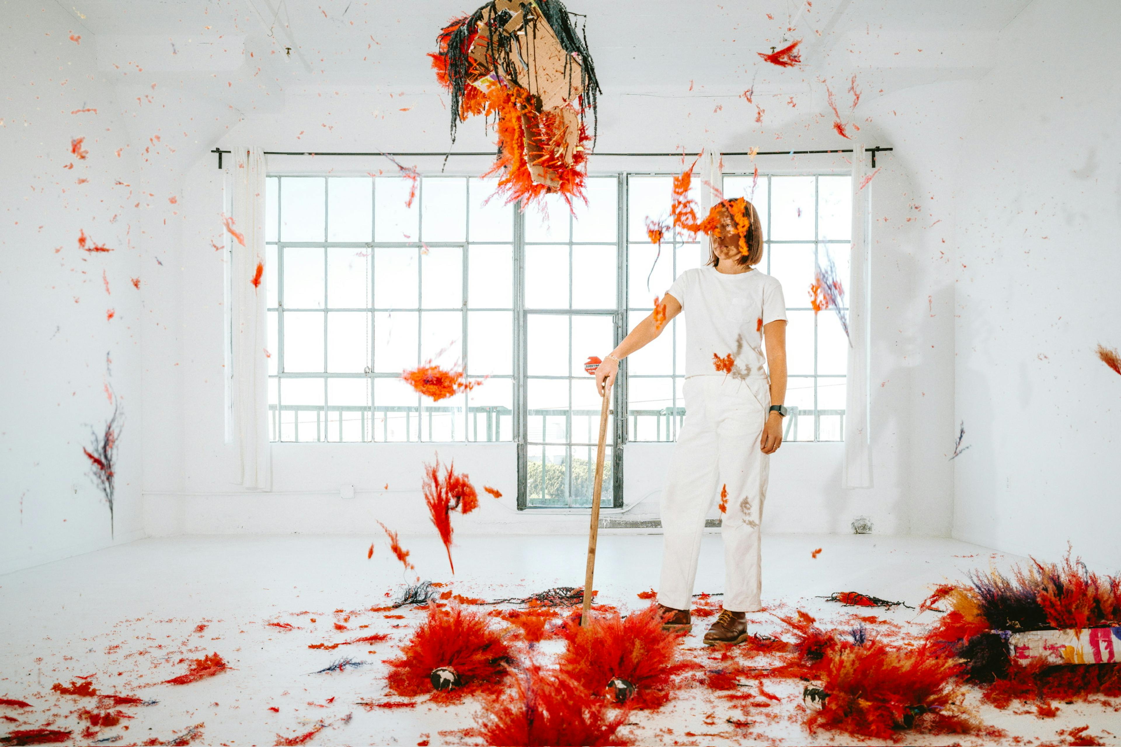 A women standing with a piñata stick behind a bright red flower-piñata that's been hit to pieces