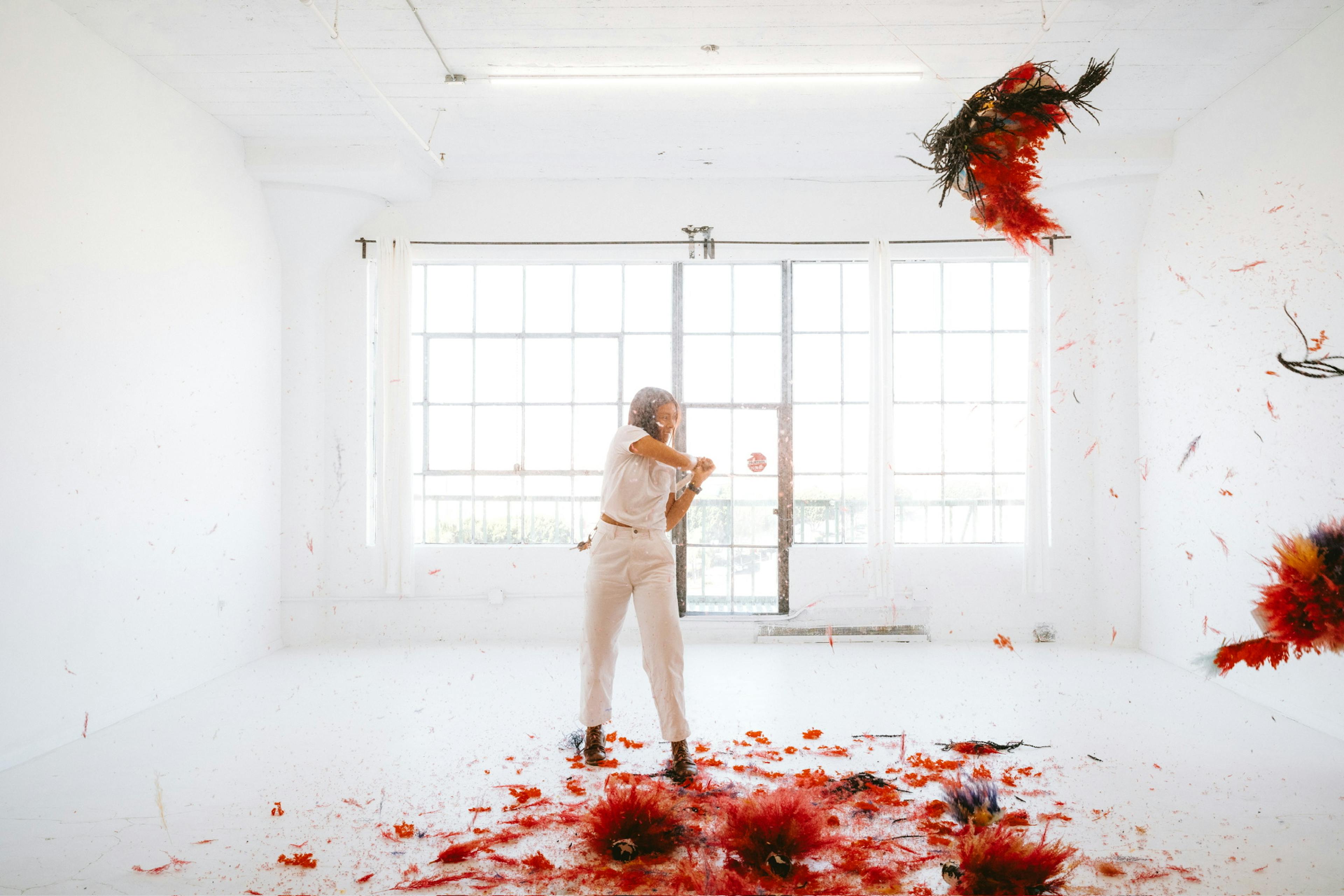 A women hitting a bright red flower-piñata with a piñata stick in a white room with a window.