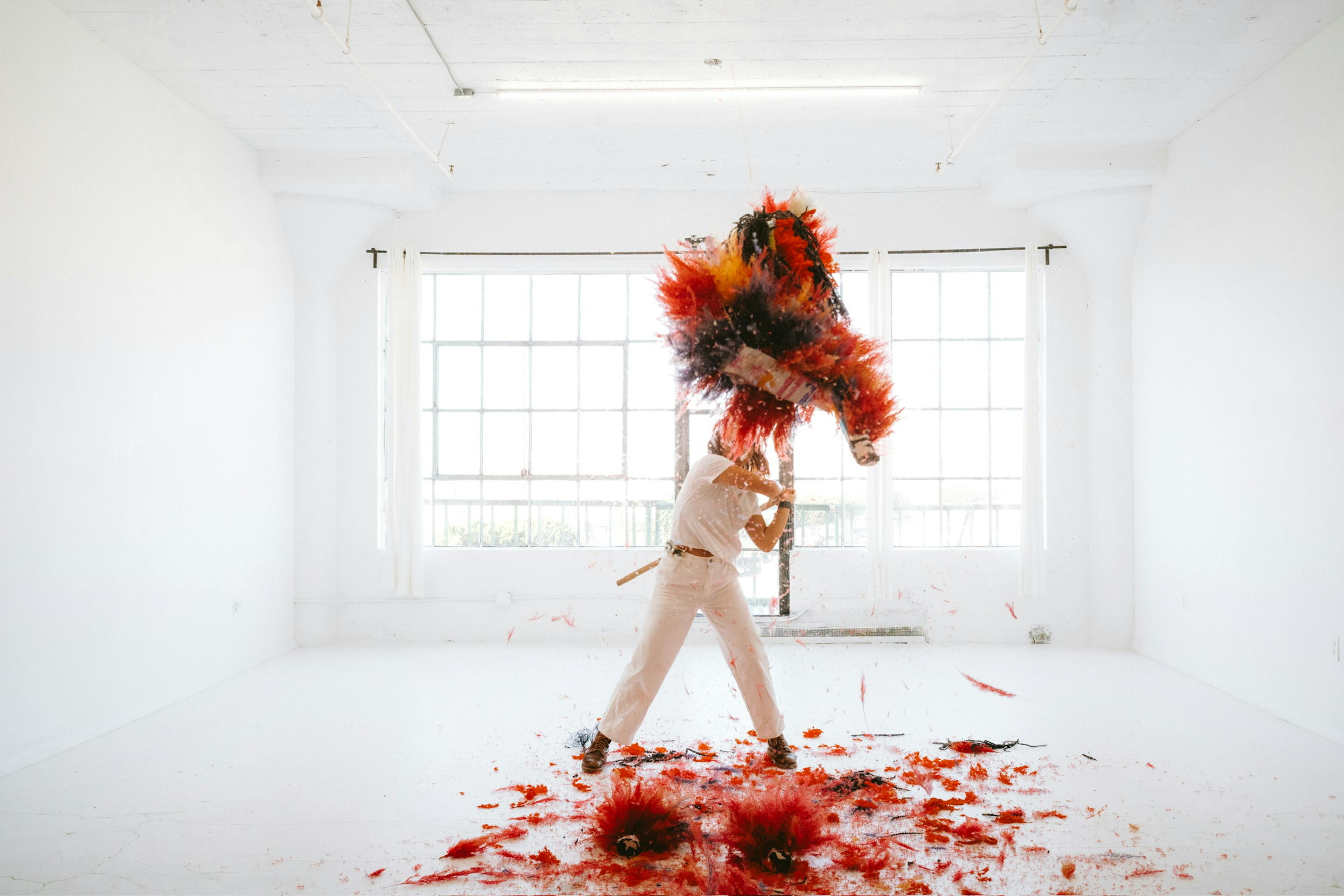 A women hitting a bright red flower-piñata with a piñata stick in a white room with a window.