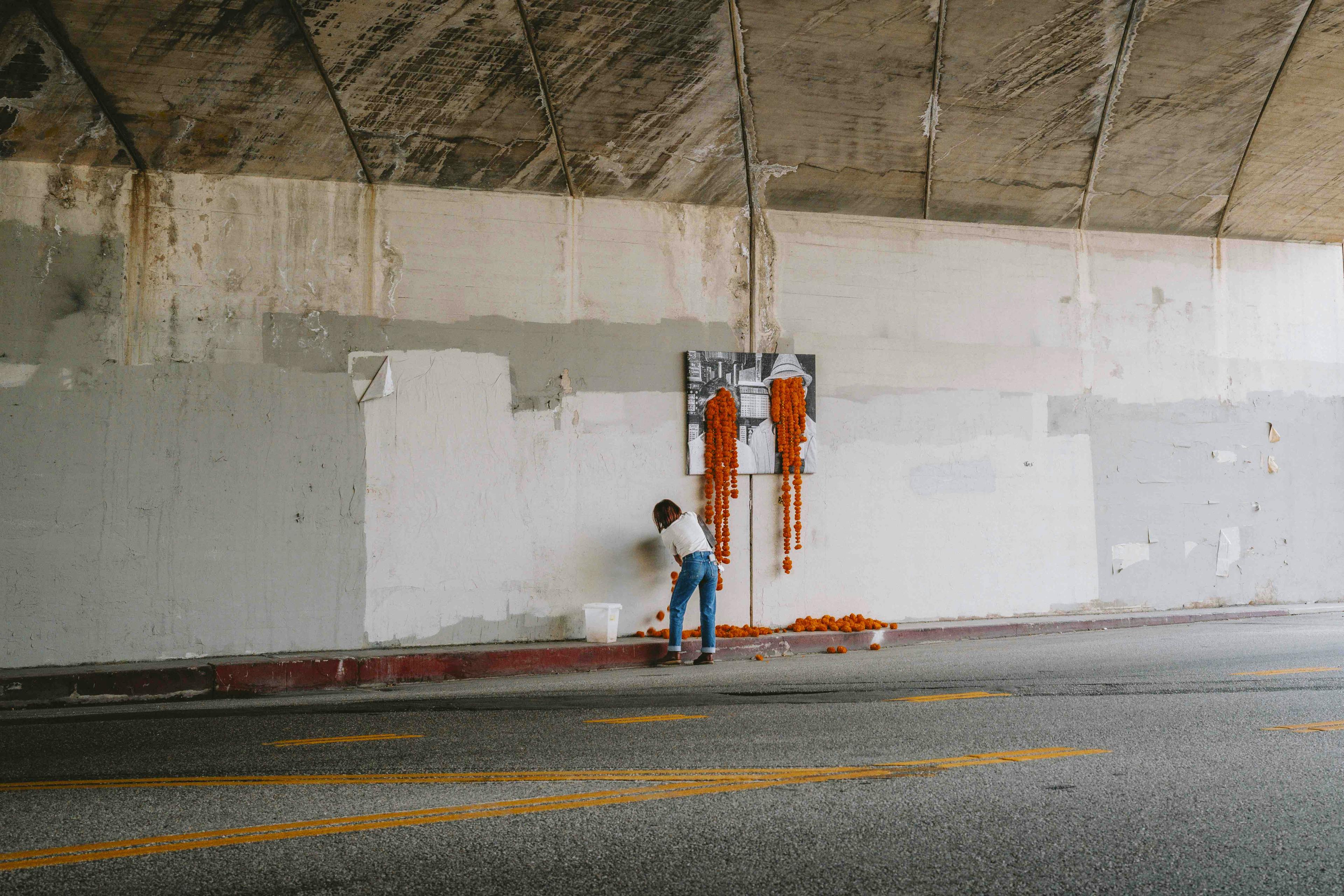 A women installing a floral art installation in an underpass with orange carnations dripping from the faces of a black and white photograph