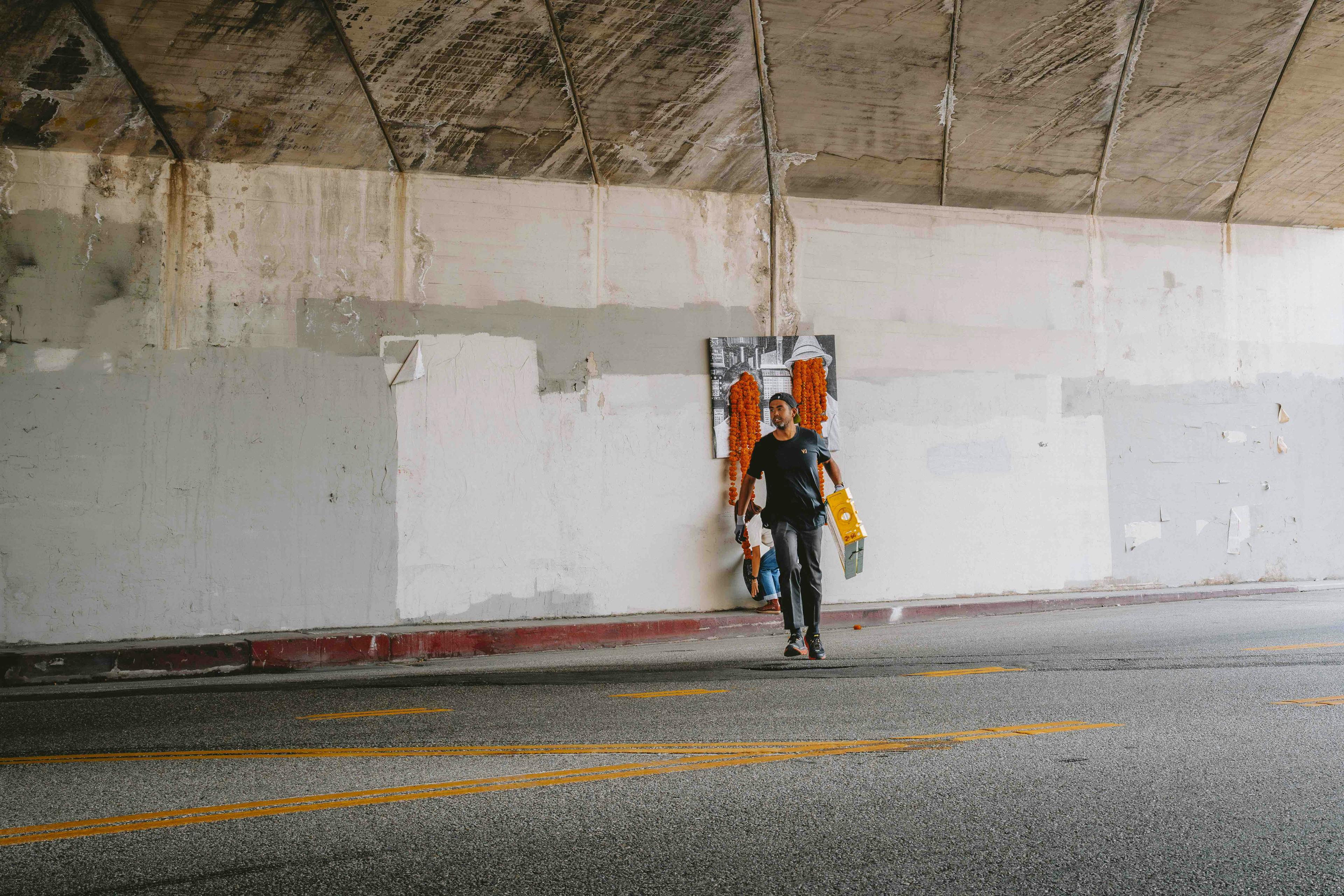 A man running with a ladder in an underpass from a floral art installation with orange carnations dripping from two faces on a black and white photograph