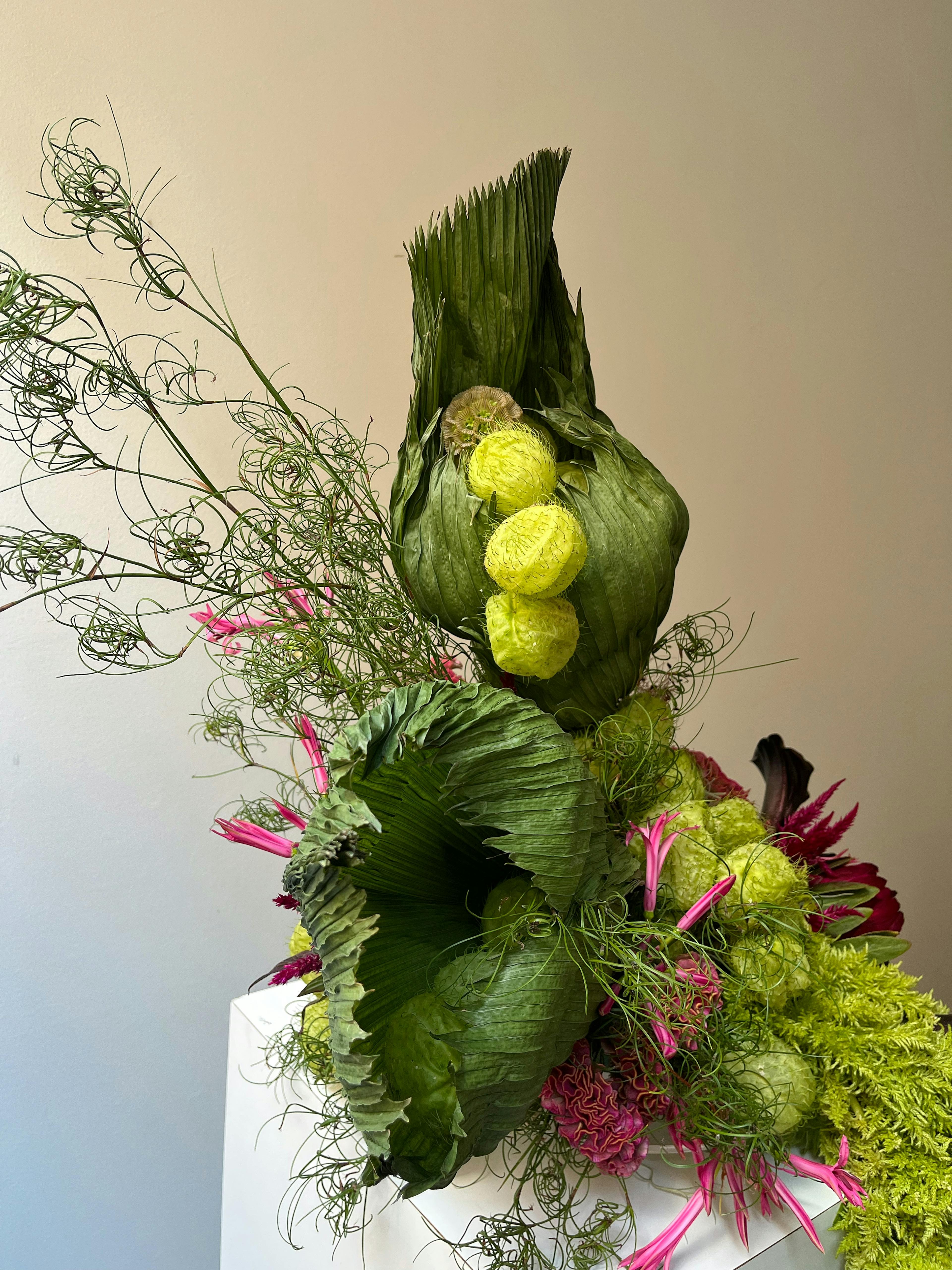 A floral arrangement on a white pedestal with puffy light and dark green plants and small pink flowers on the sides