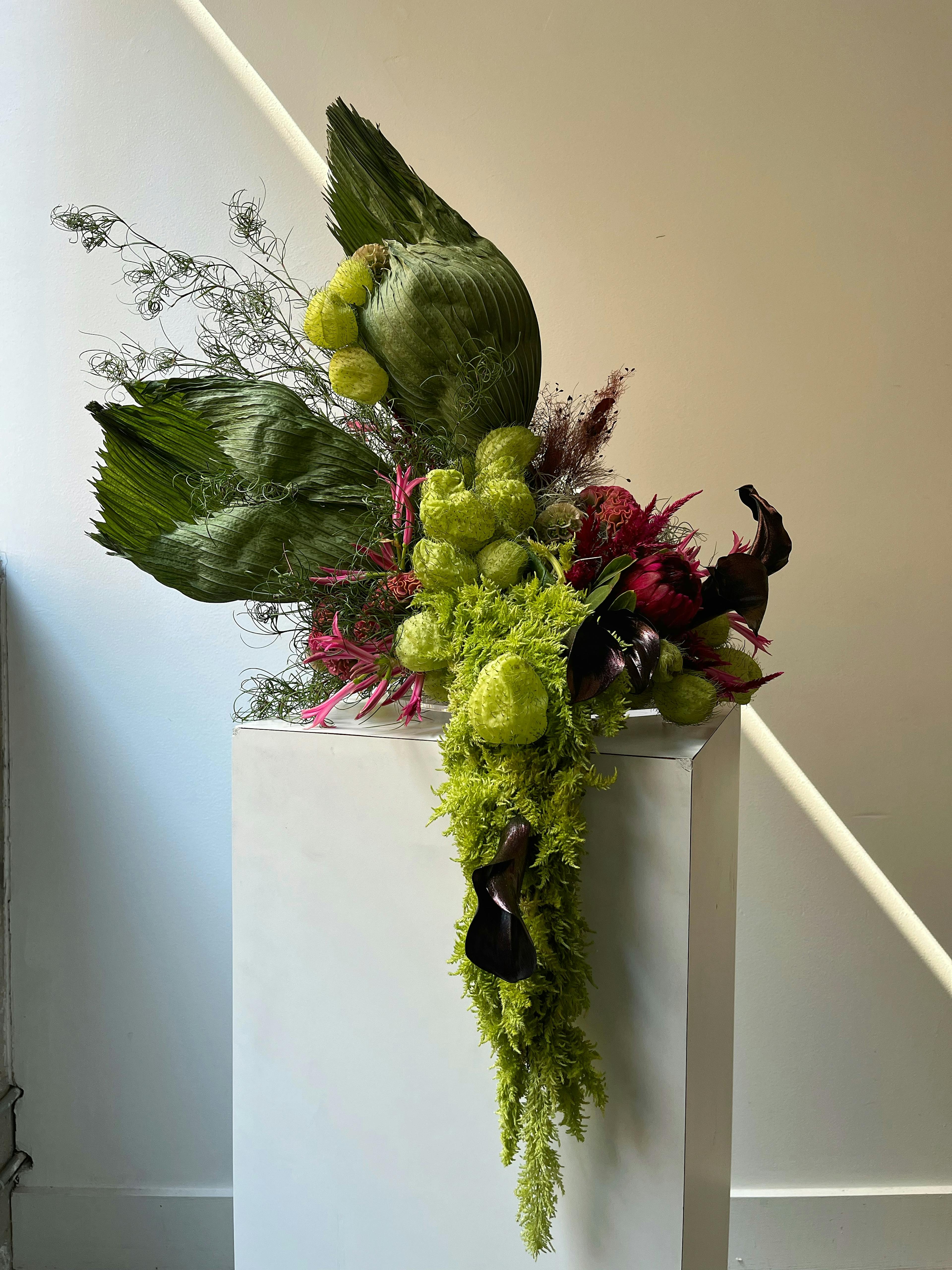 A floral arrangement on a white pedestal with puffy light and dark green plants and small pink flowers on the sides