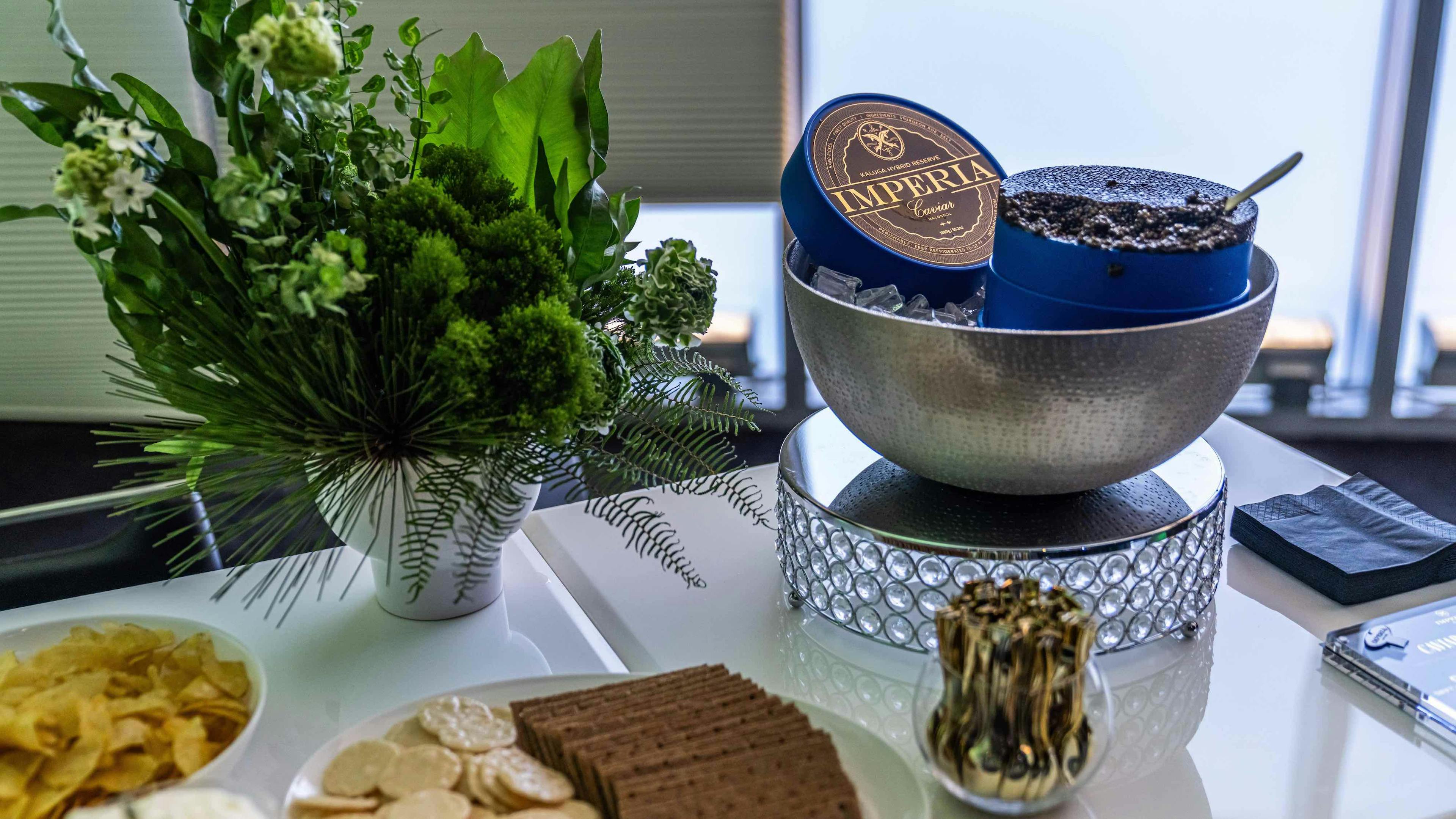 An arrangement of green foliage and white flowers with Imperia Caviar in a silver bowl on the right.