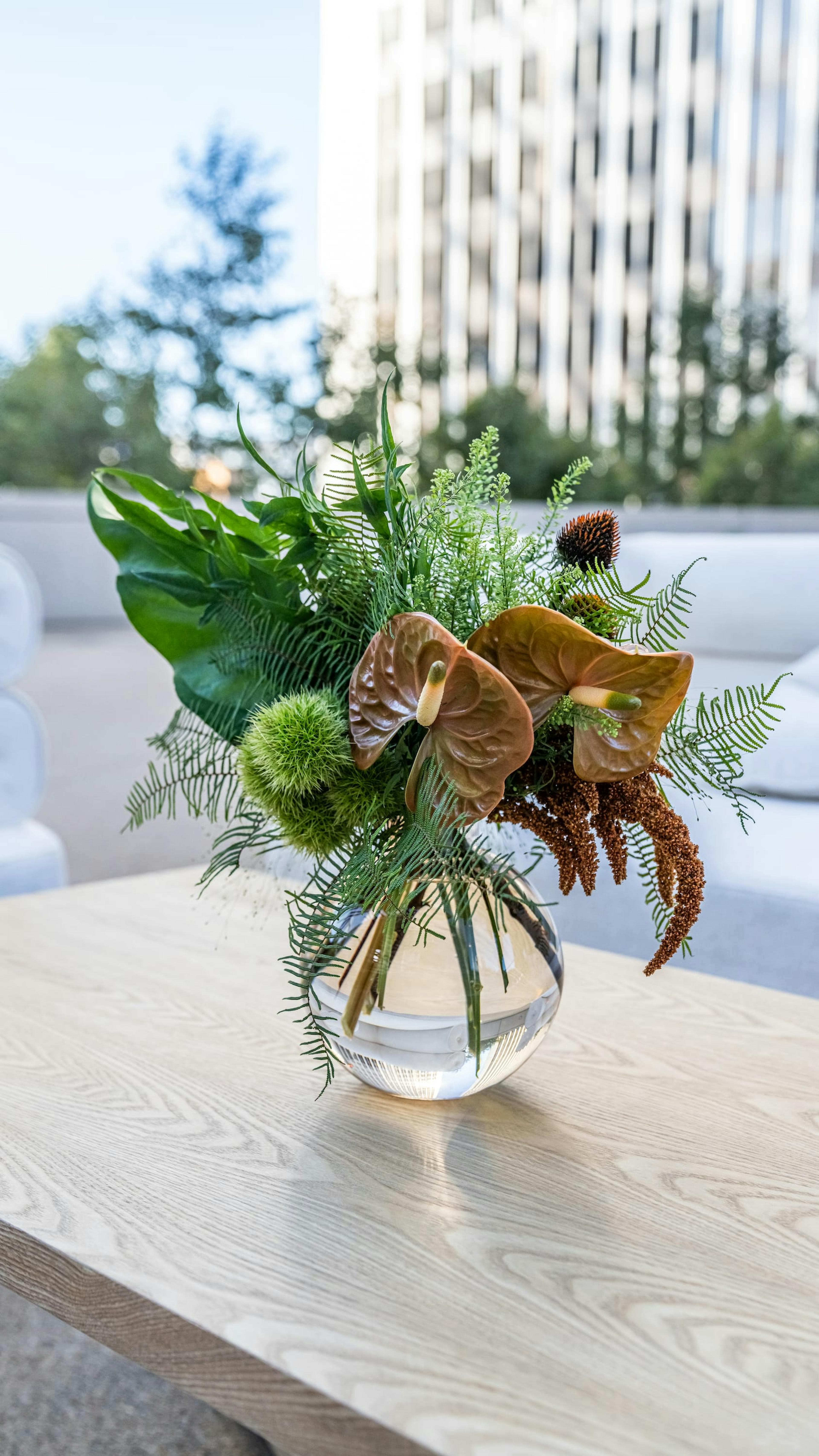 A simple arrangement of brown peace lilies, green foliage and small ferns in a clear vase on a wooden table with buildings in the background. 