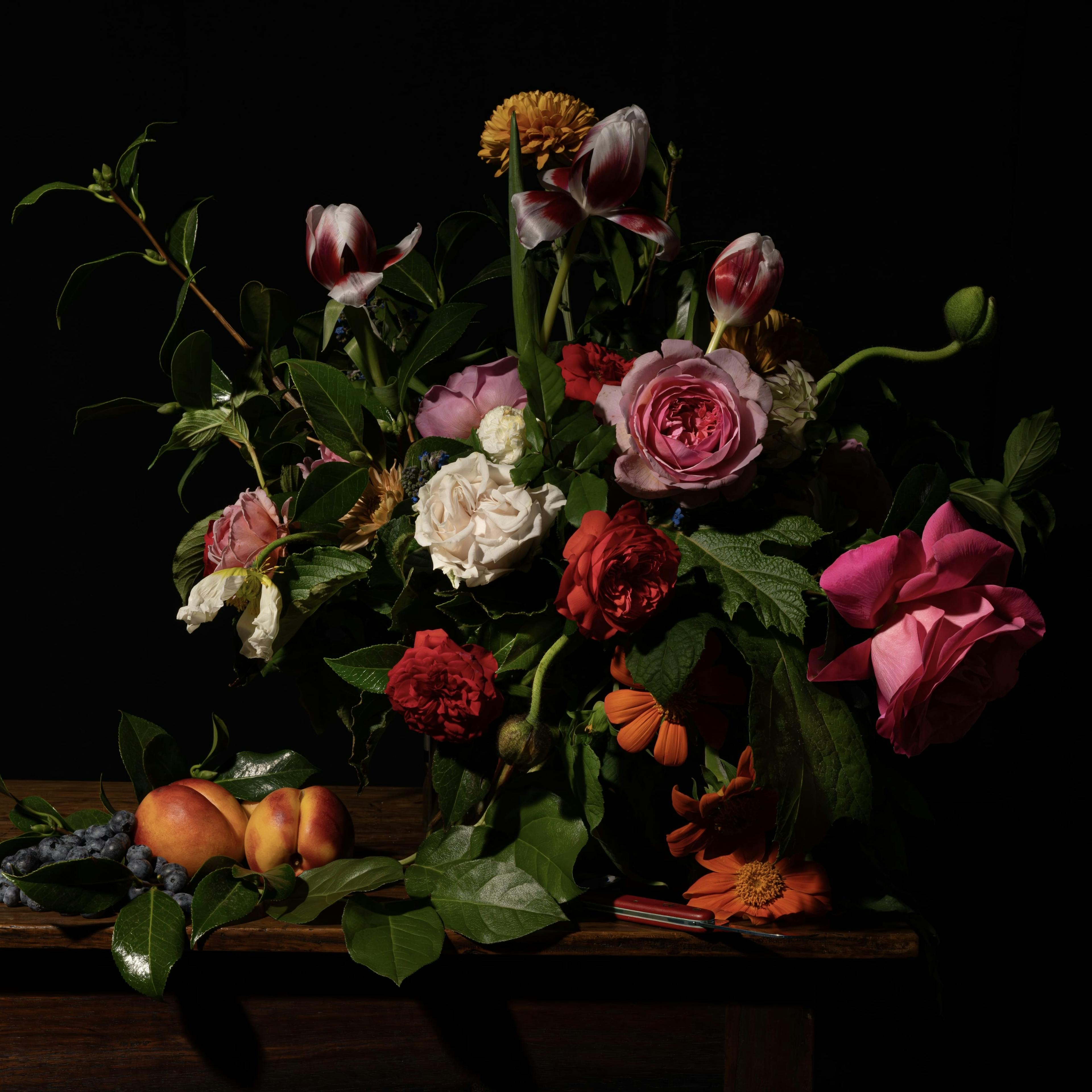 An arrangement of pink, red, white and yellow flowers and fruit in front of a black background. 