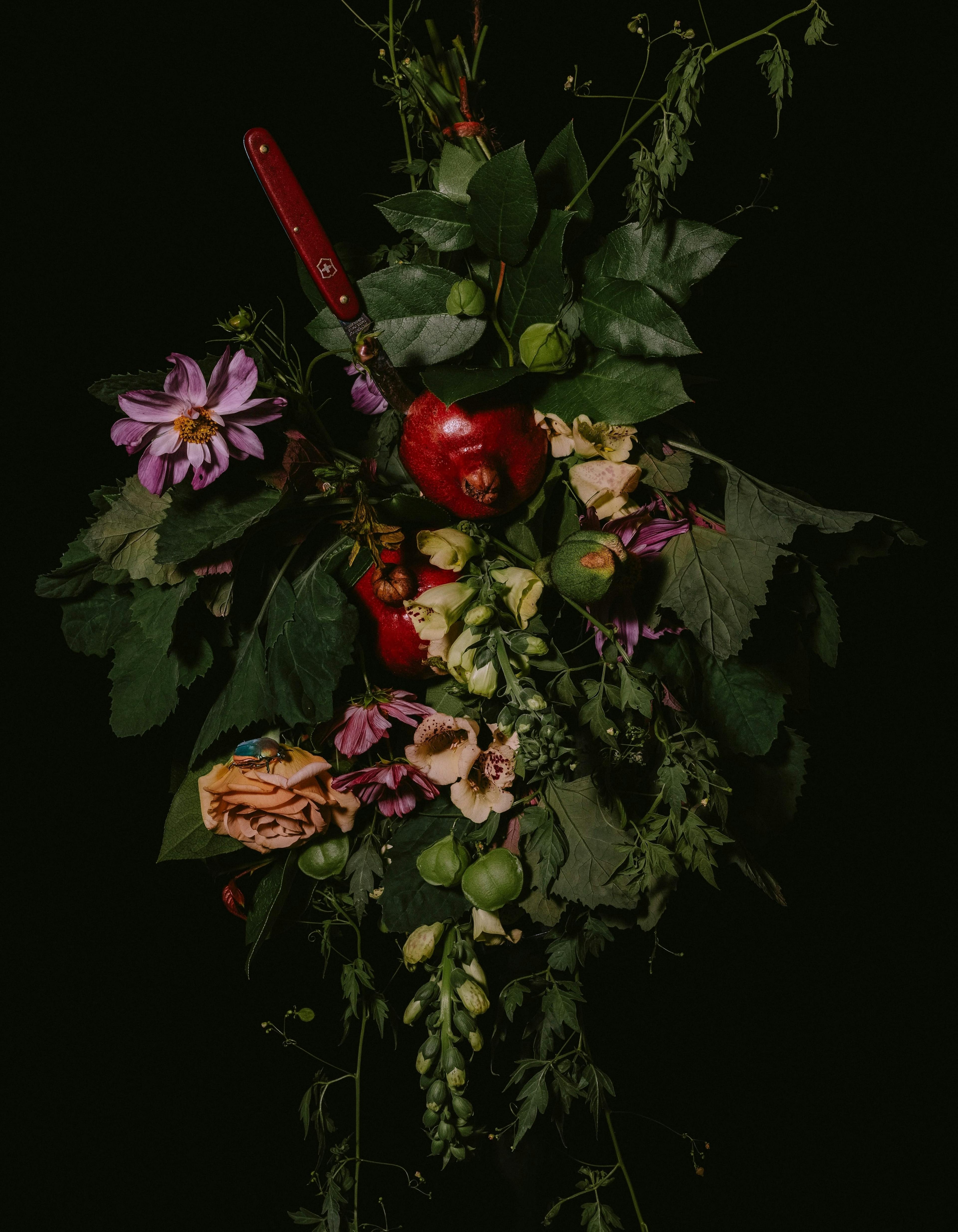 An arrangement of flowers with a pomegranate and a knife hanging on a black wall.