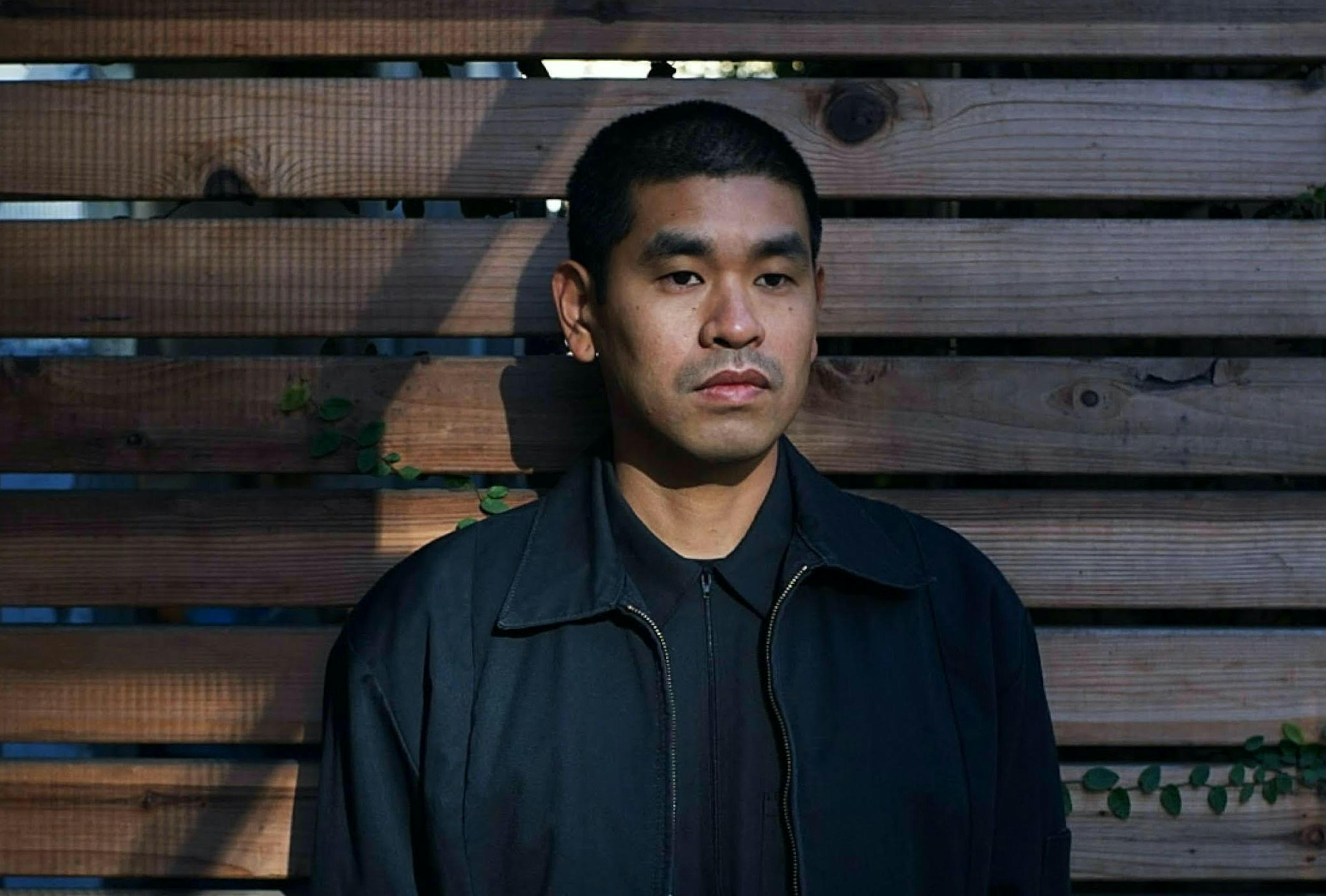 A man wearing a black jacket standing in front of a wooden slatted wall.