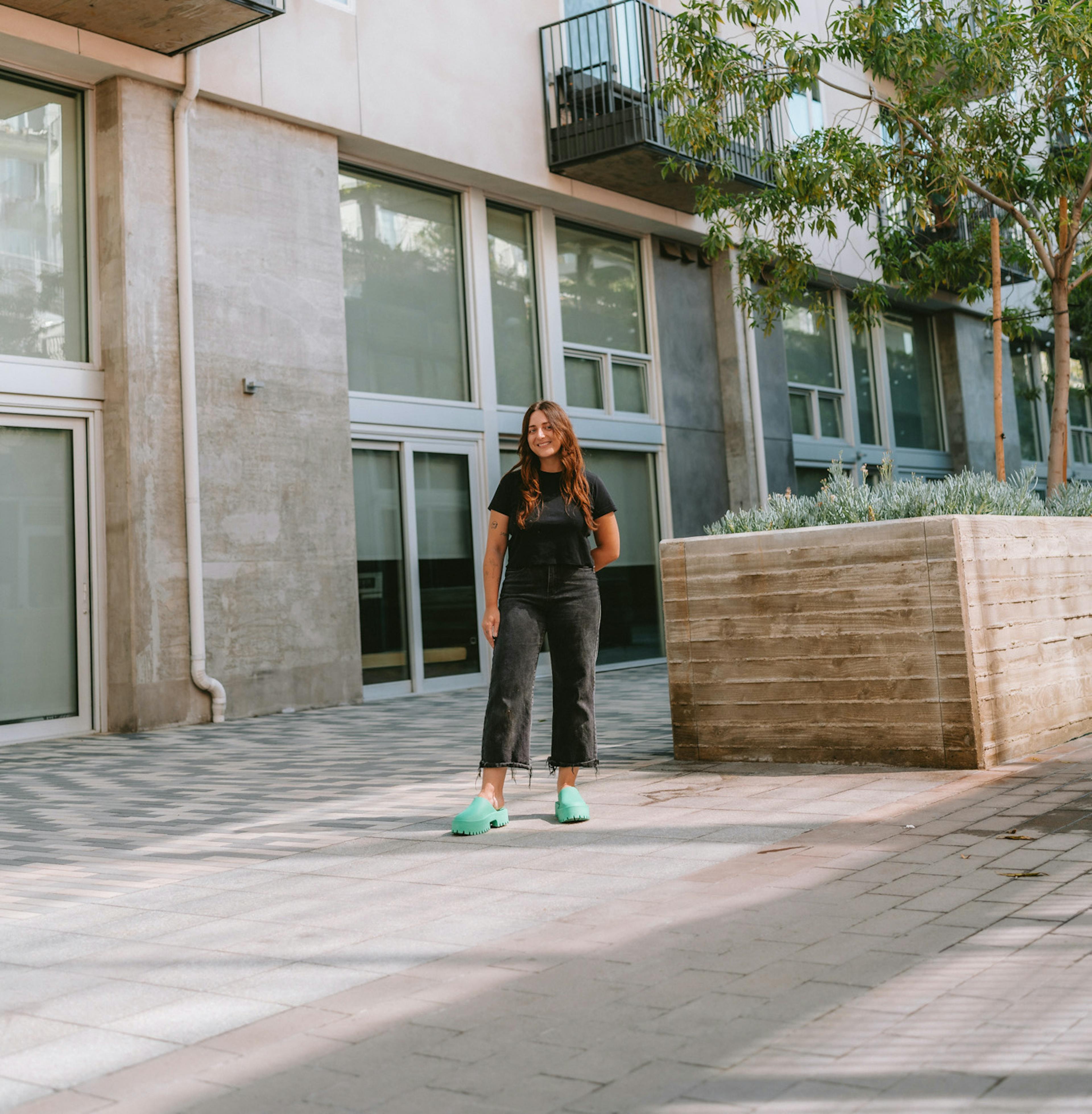 A girl wearing all black clothes and bright blue platform crocs standing in an urban landscape