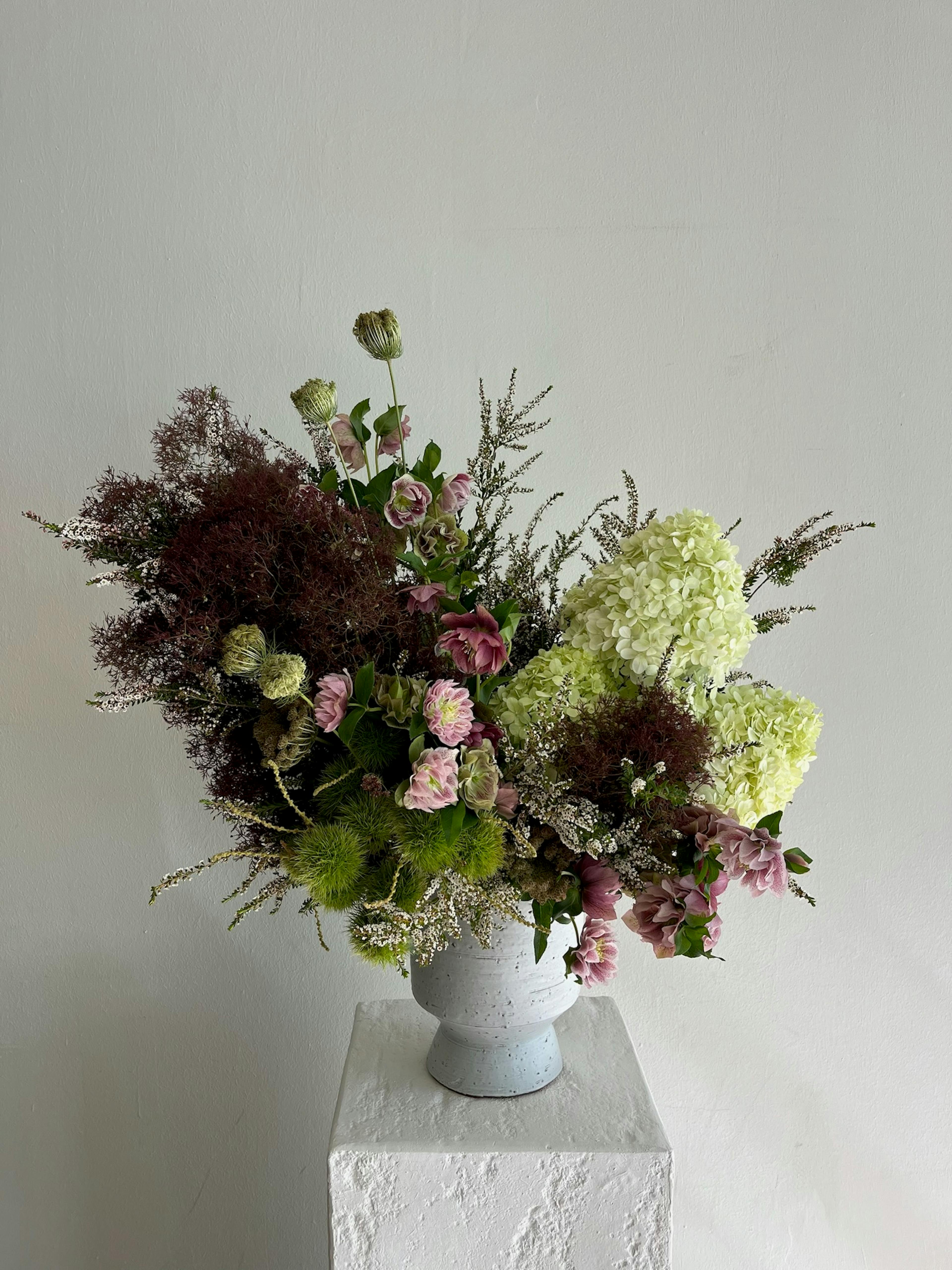 An arrangement of light green, dark purple and pink flowers on a pedestal in front of a white wall