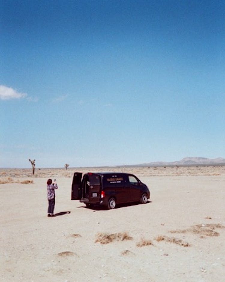 A far-away shot of a women taking photos of a black van in the desert.