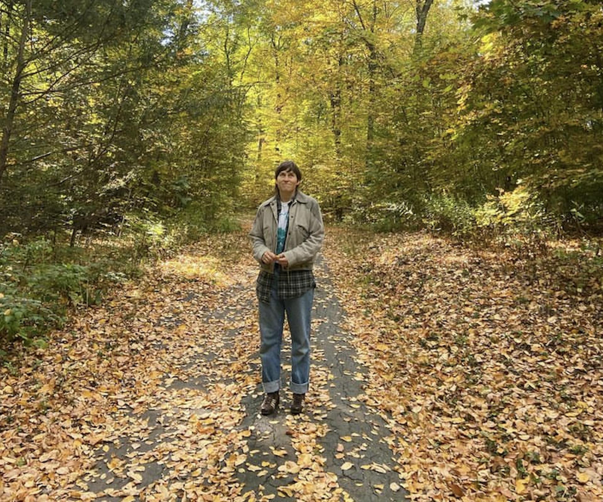 A person with short hair wearing a tan jacket standing in the woods in fall